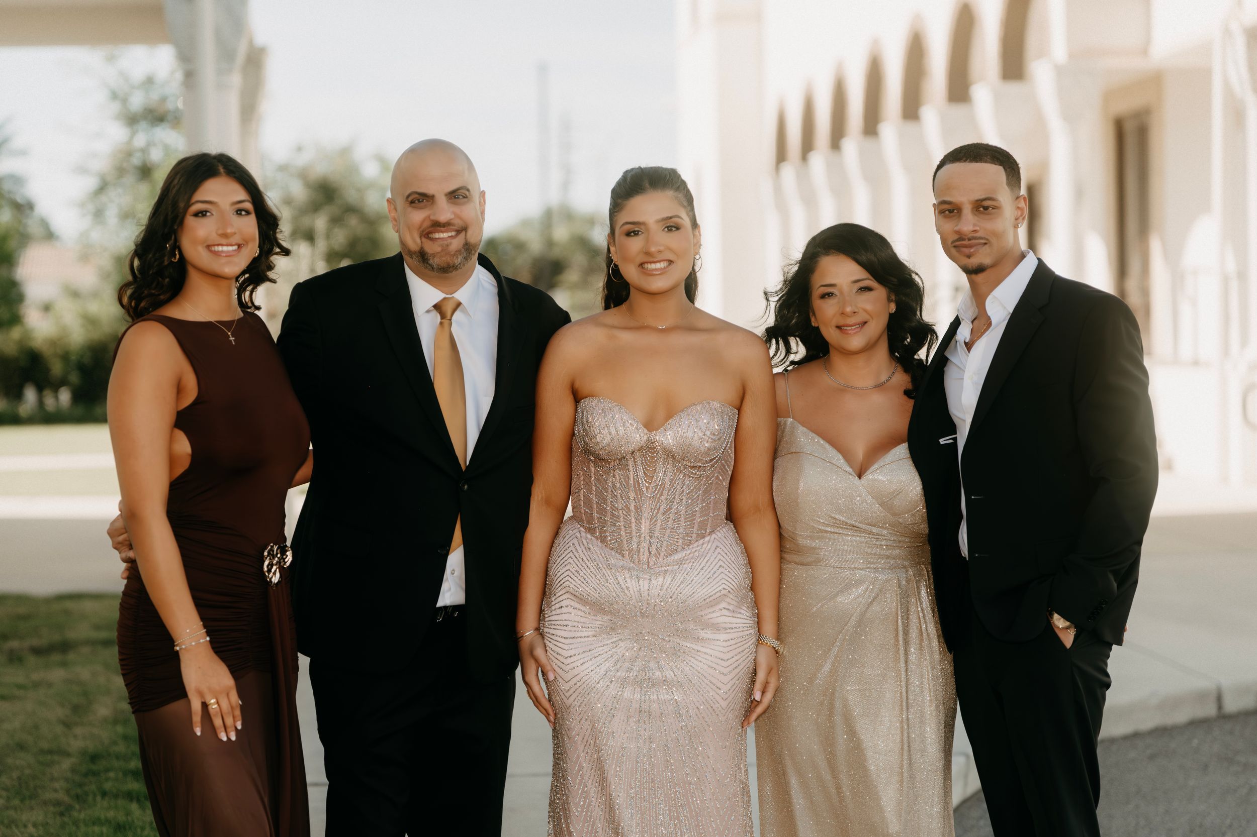 A group of five elegantly dressed people posing together at a formal wedding celebration outdoors.