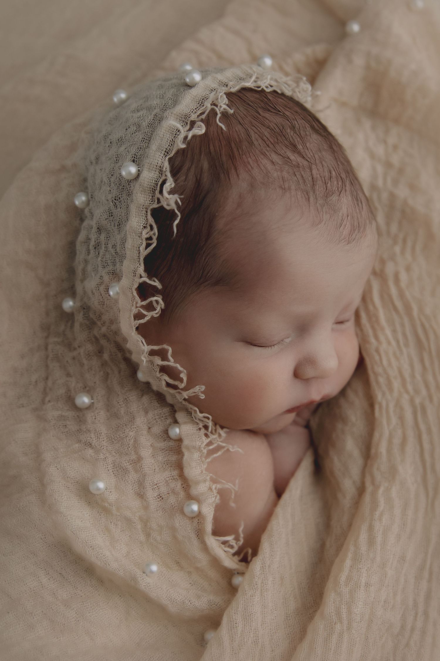 Peaceful sleeping newborn wrapped in soft beige fabric with delicate pearl details and lace trim.
