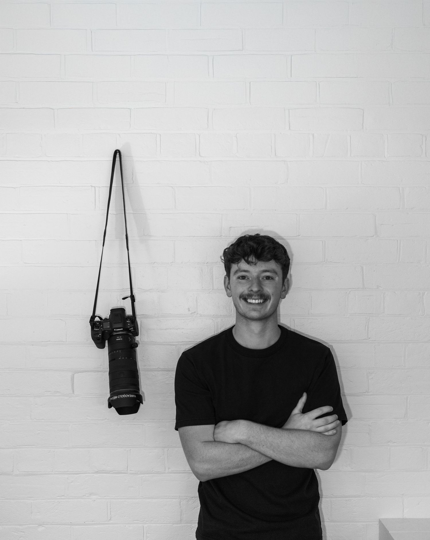 Black and white portrait of photographer Jayden Threadgold standing against a brick wall with a camera hanging nearby for Threadgold Media