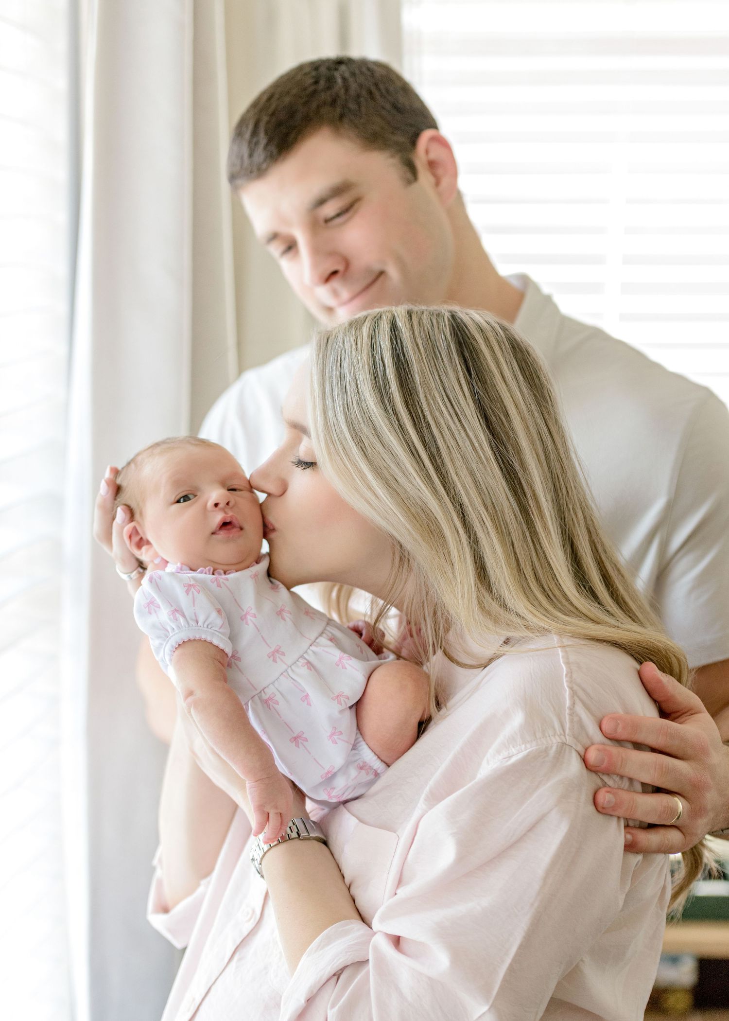 Mom kissing newborn daughters cheek as baby girl looks at camera, and dad admires them both from behind.