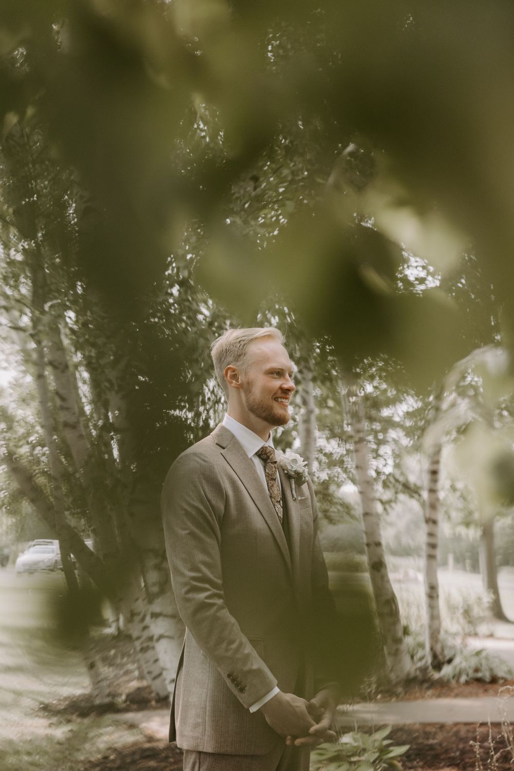 Smiling groom in a tan suit with floral tie standing among birch trees outdoors.