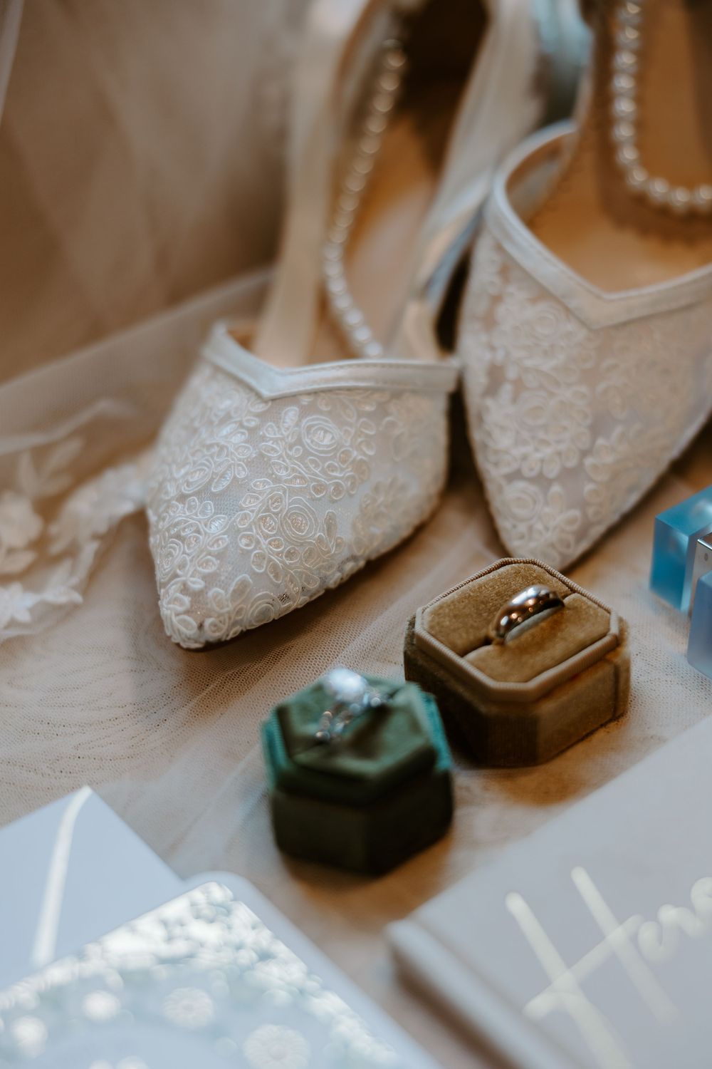 Lace bridal heels with pearl anklets displayed alongside wedding rings in green velvet boxes on a wooden surface.