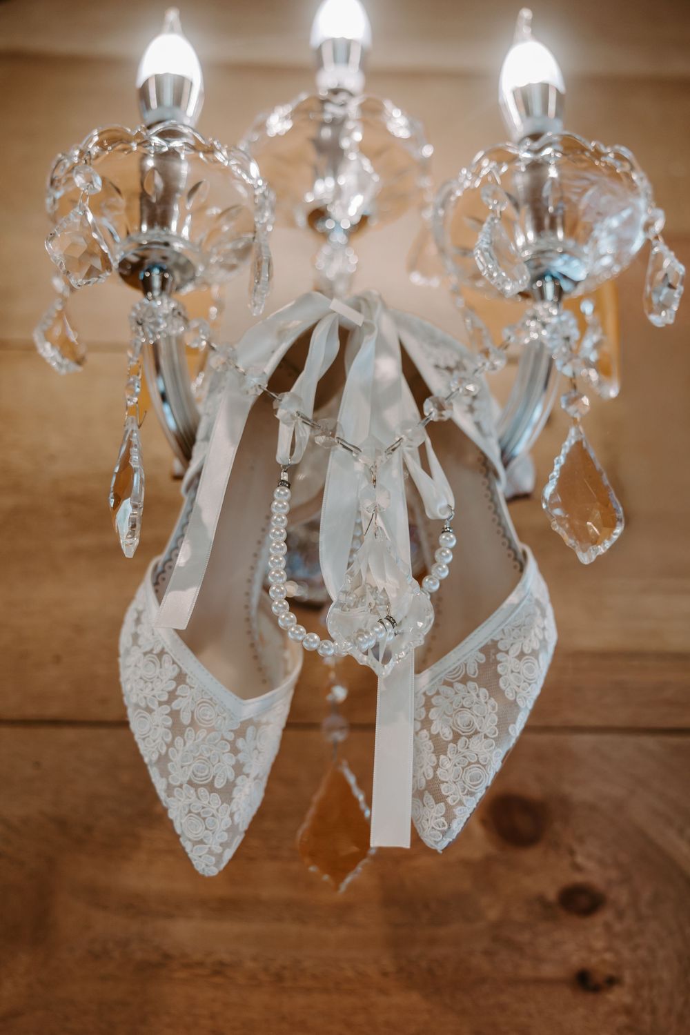 White lace bridal heels with ribbon ties hanging elegantly from a crystal chandelier.