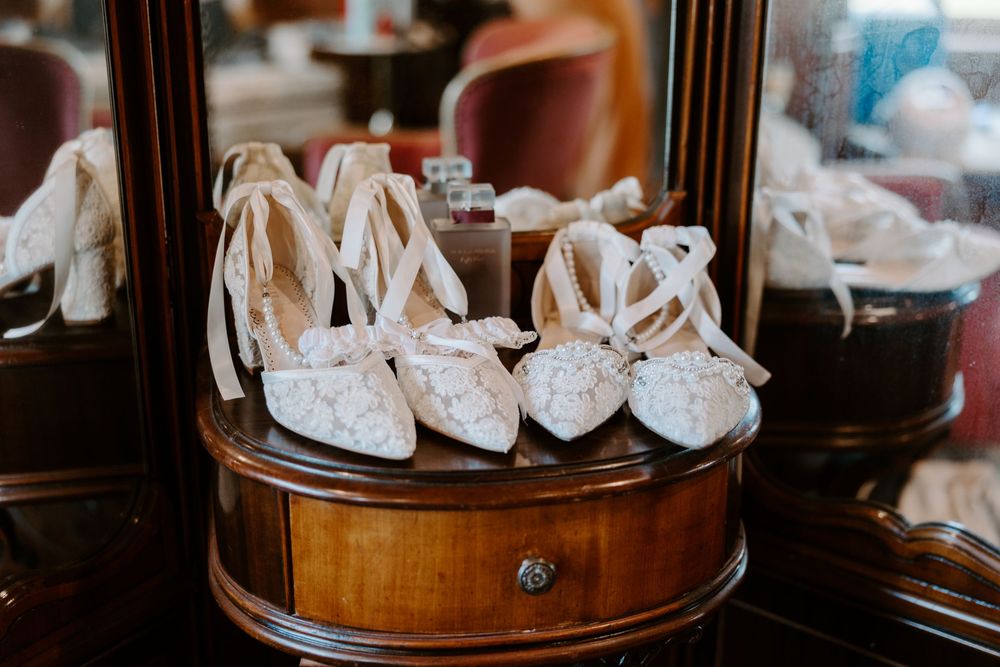 Two pairs of white lace bridal heels with ribbon ankle ties displayed on a wooden dresser.