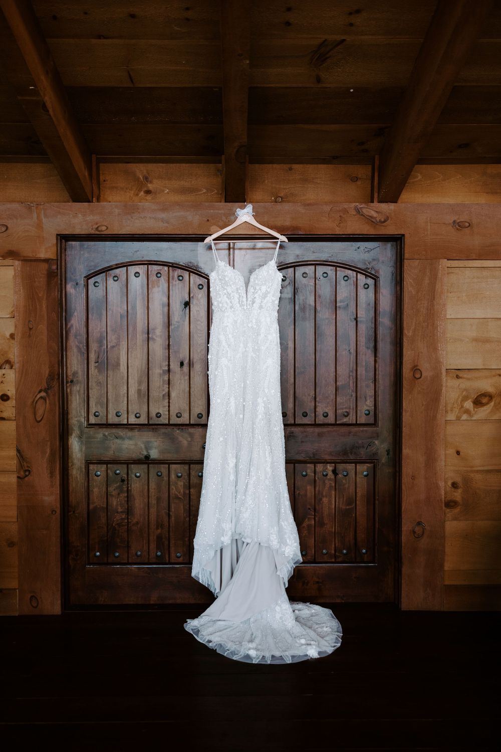 White lace wedding gown with train hanging on a hanger against rustic wooden barn doors.