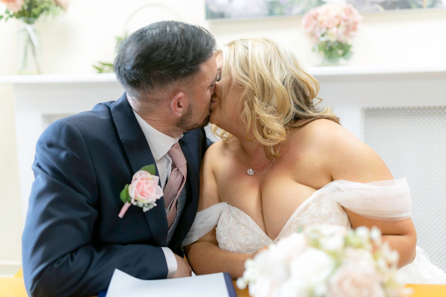 A bride and groom share a romantic kiss during their wedding ceremony, with the groom in a navy suit and pink boutonniere.
