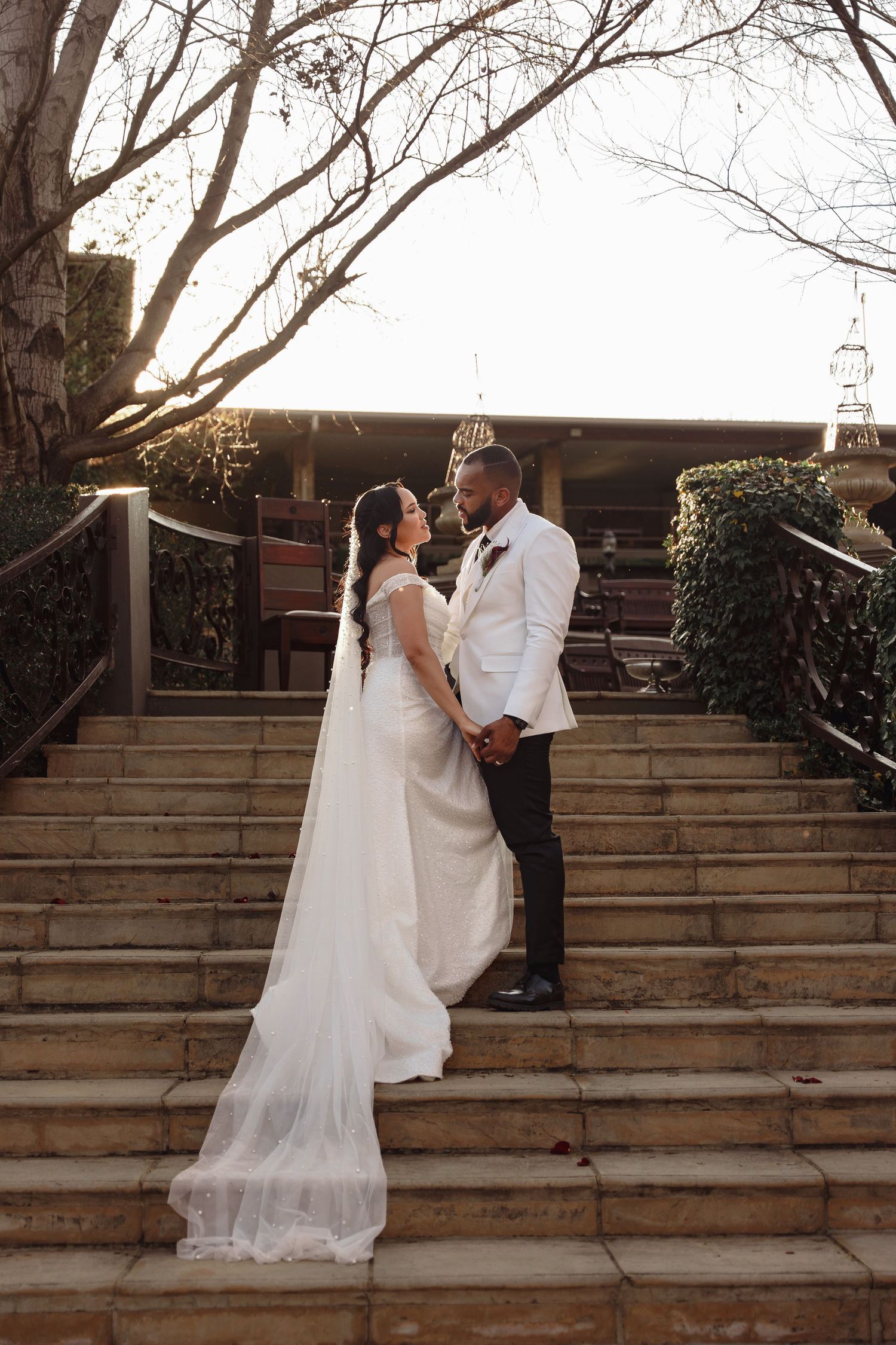 Bride and groom stand on stone steps holding hands, bride in white gown with veil, groom in white jacket.