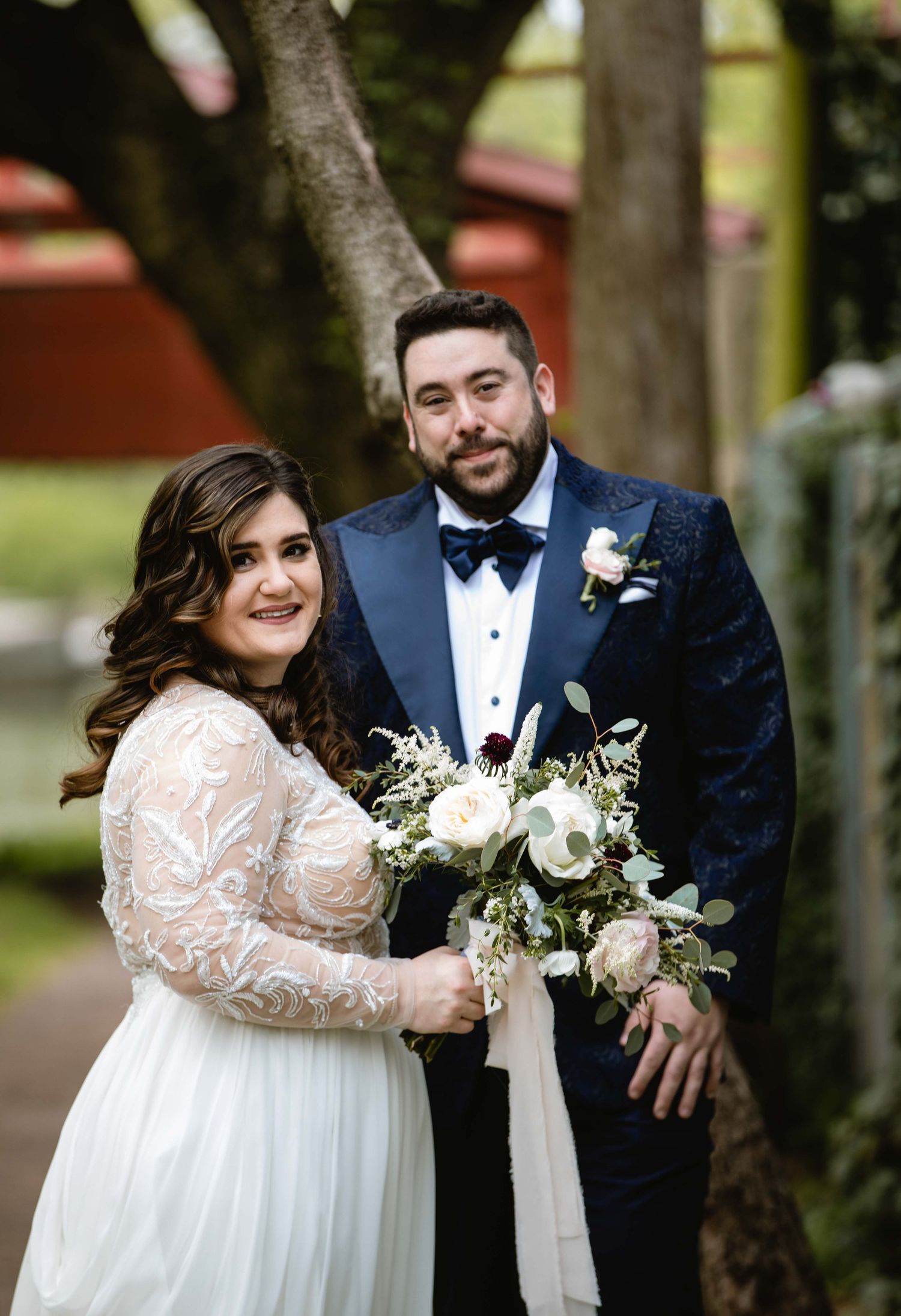 A romantic wedding couple in navy tuxedo and white lace dress pose together outdoors among trees.