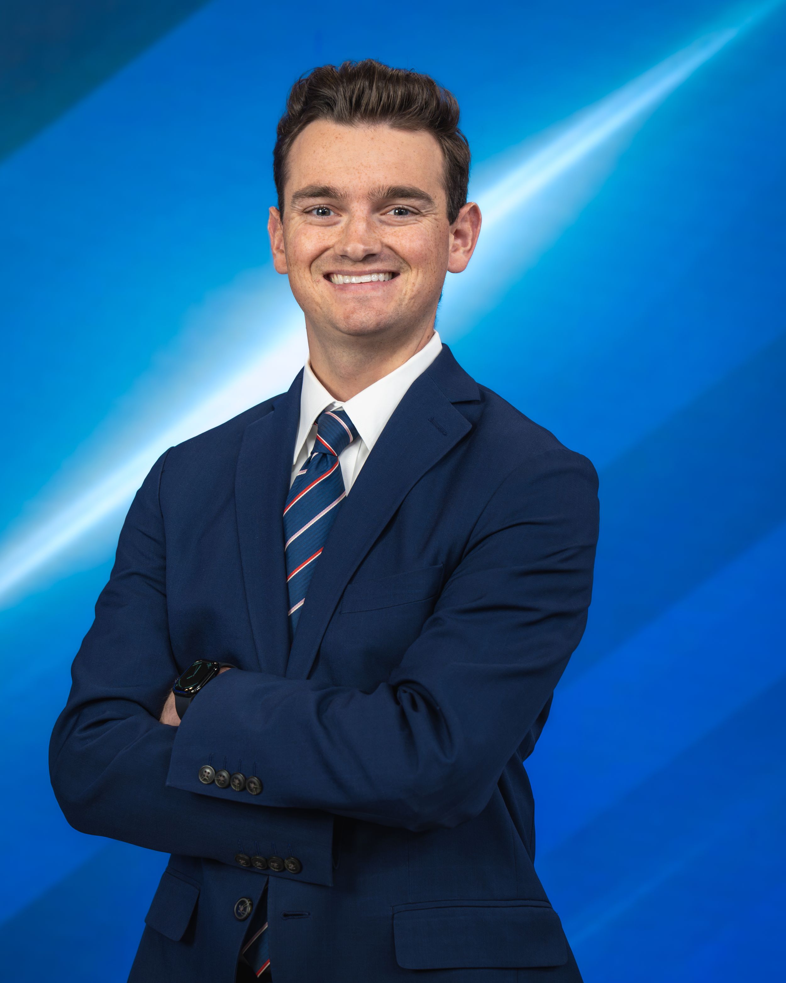 Young professional man in navy suit and striped tie smiling with arms crossed against blue background.