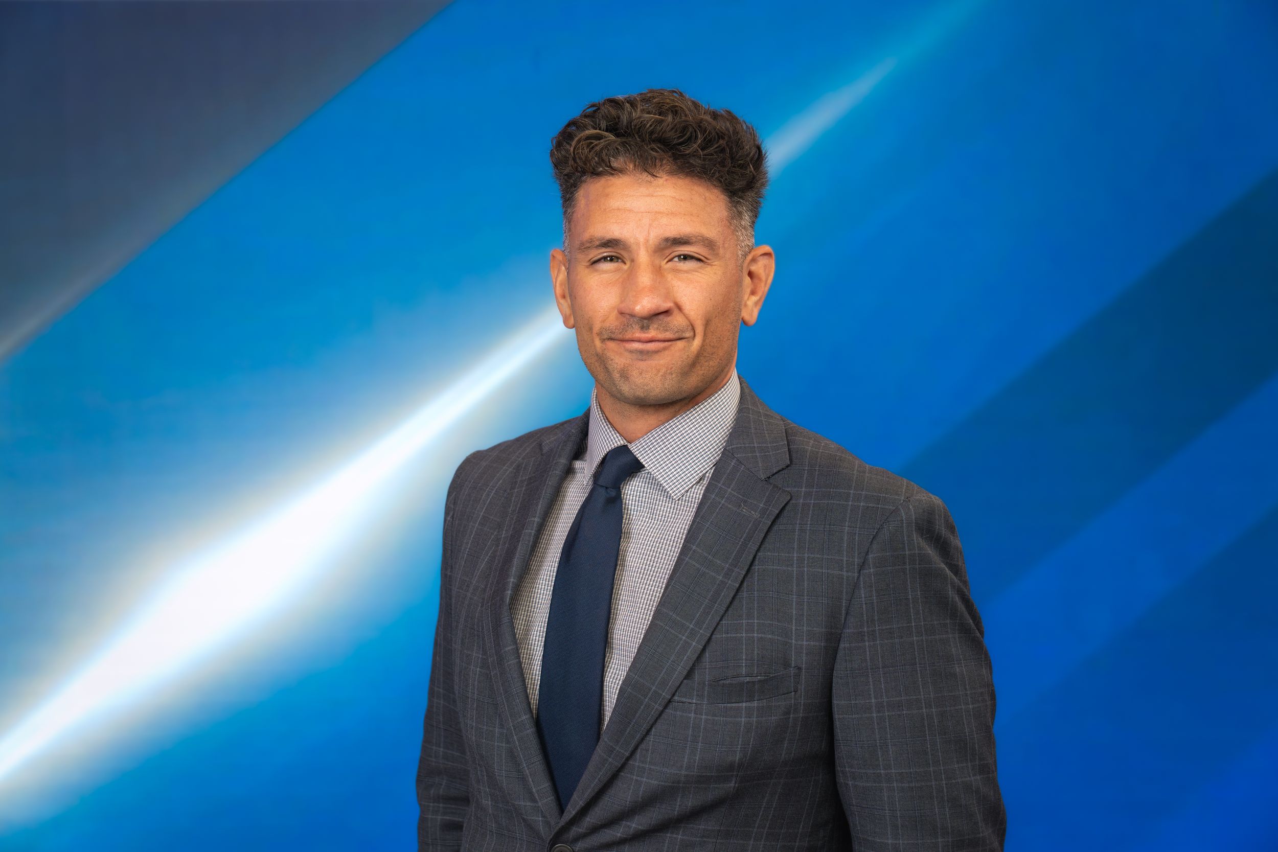 Professional man in gray suit and navy tie poses against a blue background for a corporate headshot.