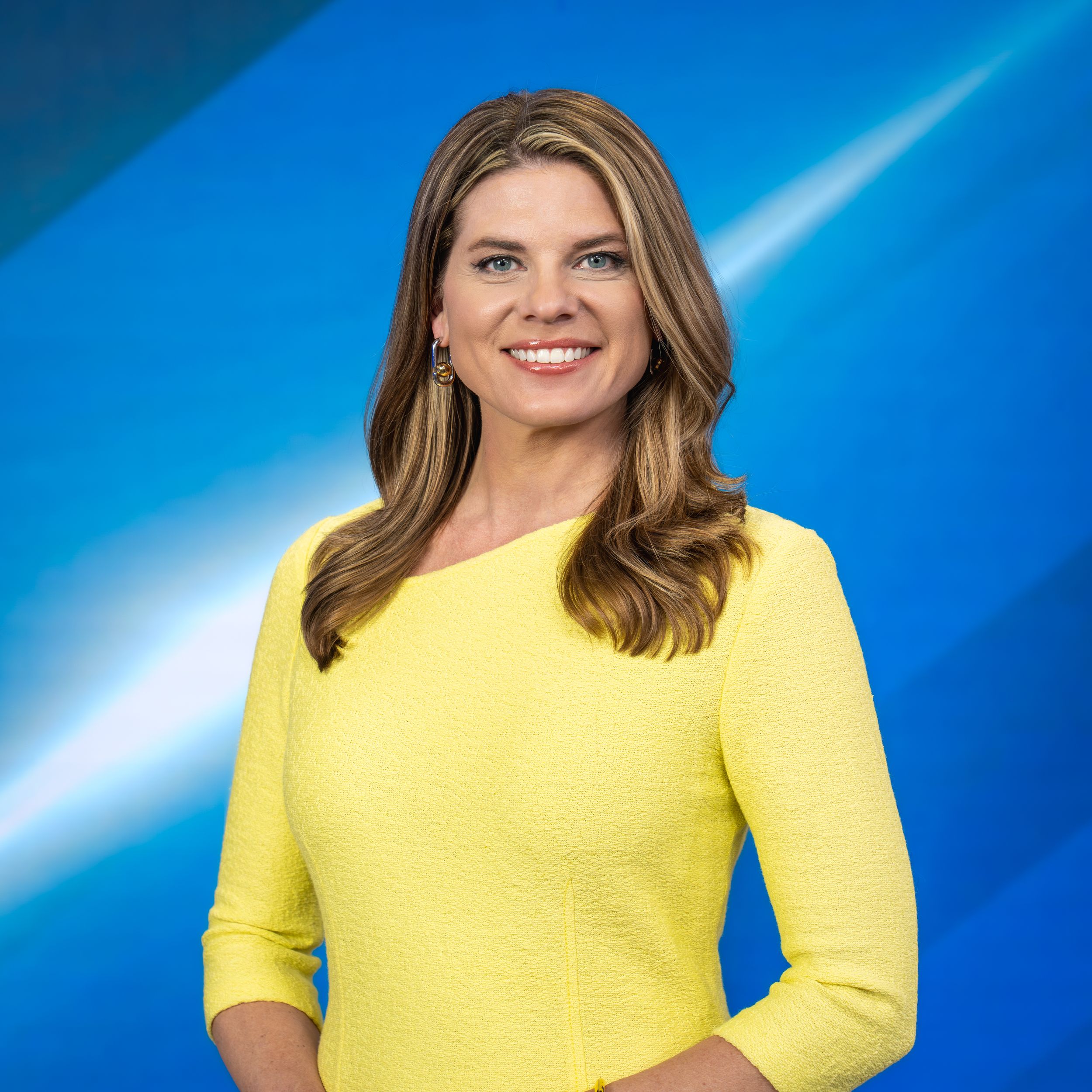 Professional woman in yellow dress smiling confidently against a blue background, ideal for news anchor or media profile.