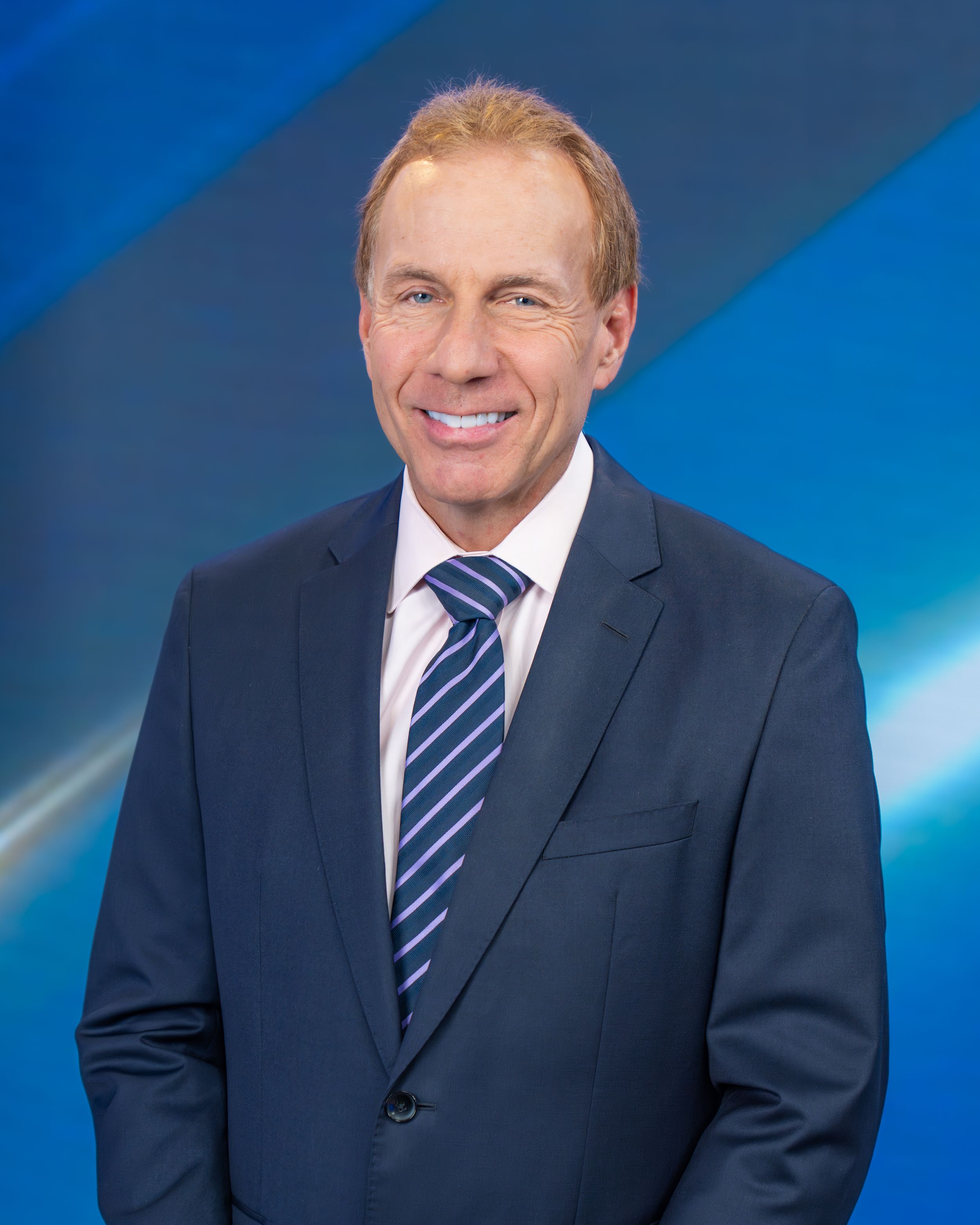 Professional headshot of someone in a navy suit and striped tie against a blue gradient background.