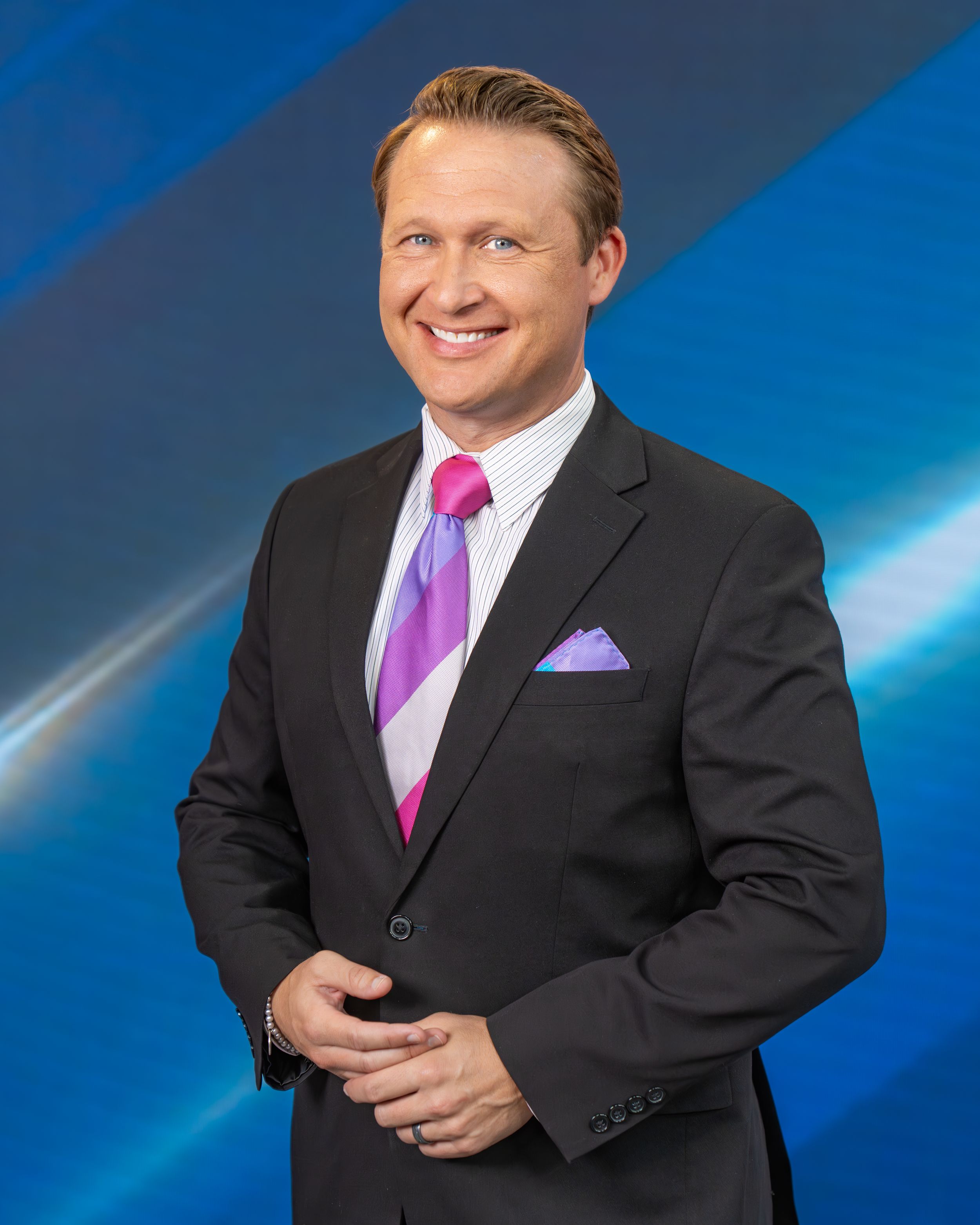 Professional headshot of someone in a dark suit with purple tie and pocket square against a blue studio background.