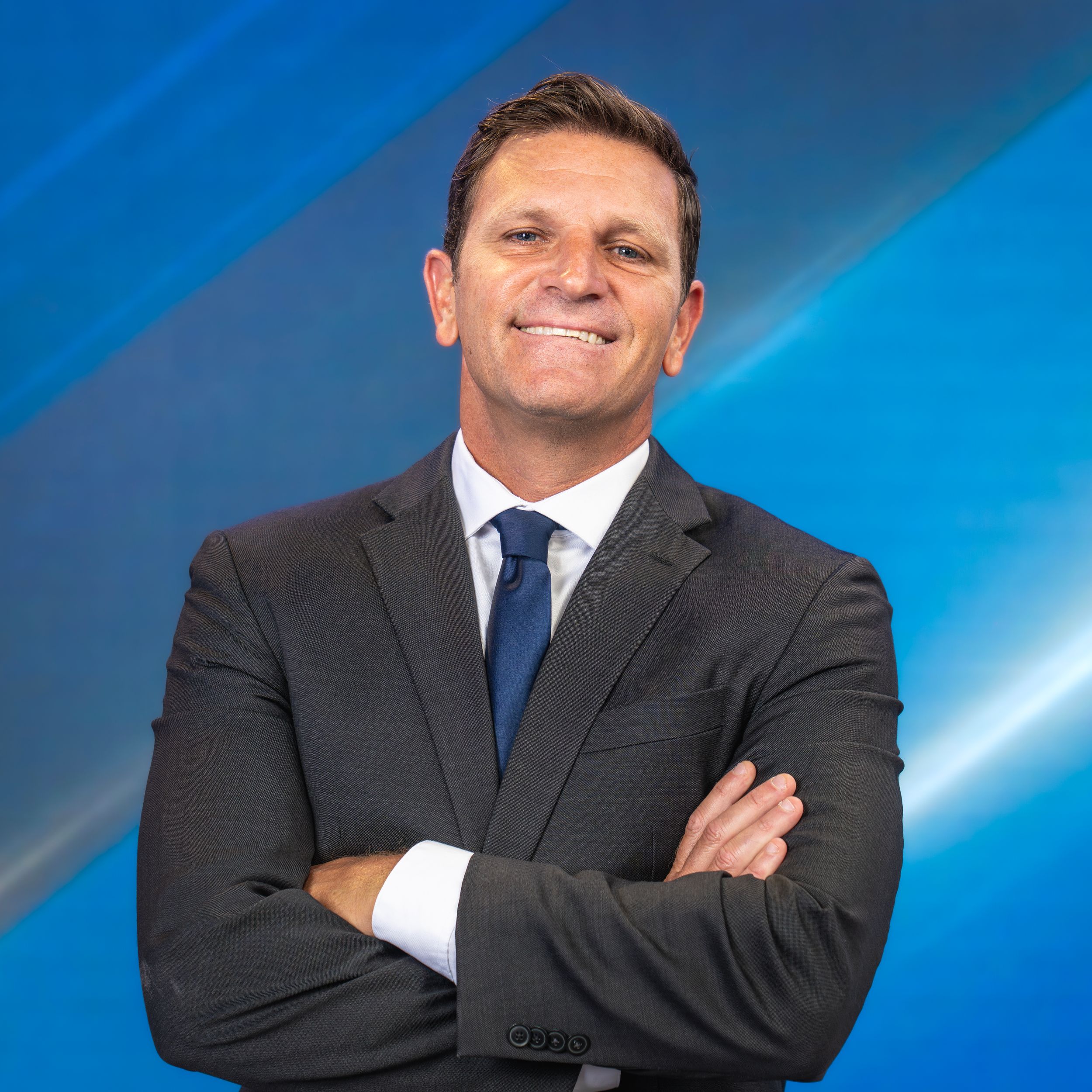 Professional portrait of person in dark suit and blue tie against blue background smiling with arms crossed.