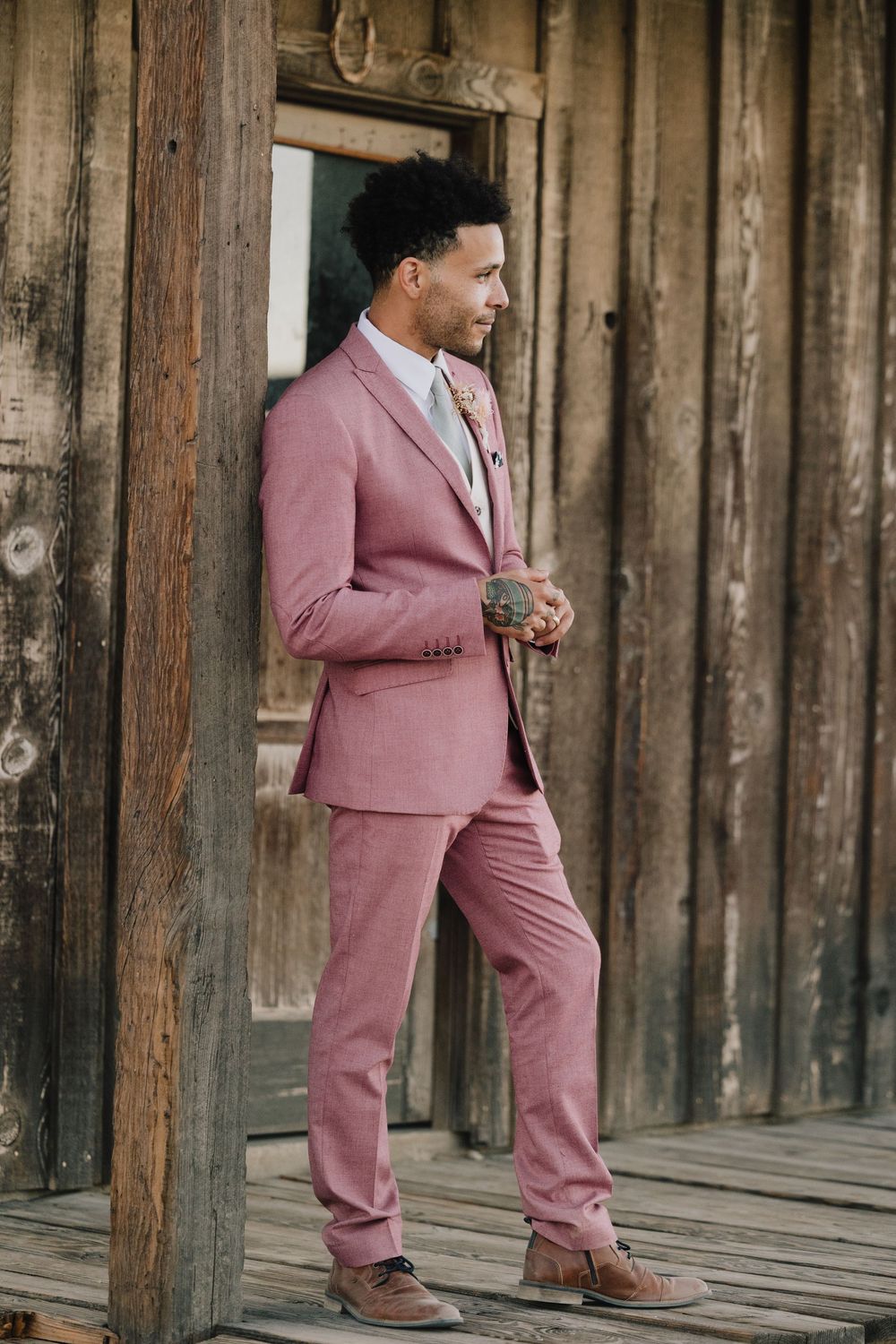 Groom in pink suit leans against rustic wooden building, looking away thoughtfully.