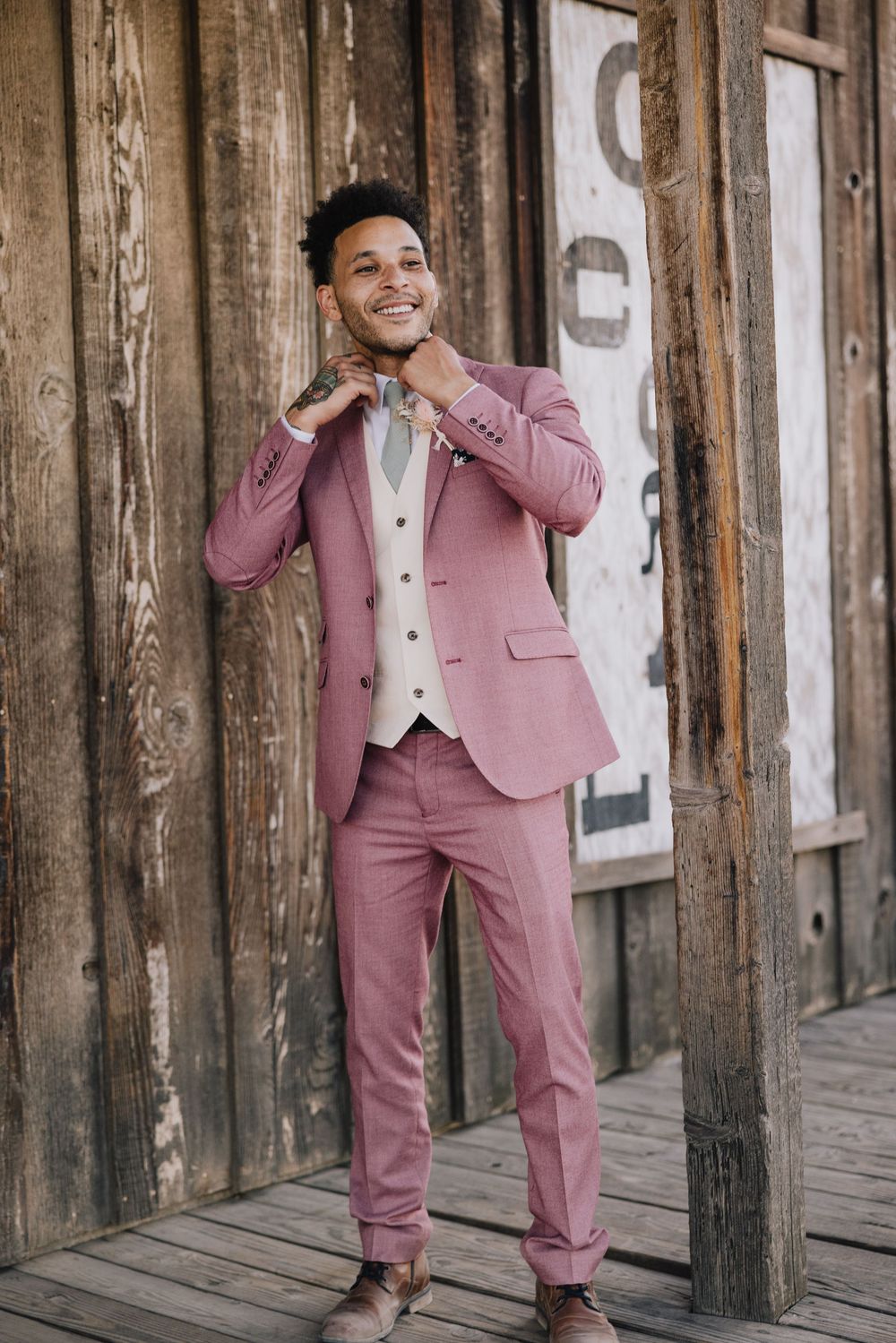 Stylish groom in pink suit and vest adjusts bow tie while smiling against rustic wooden barn wall.