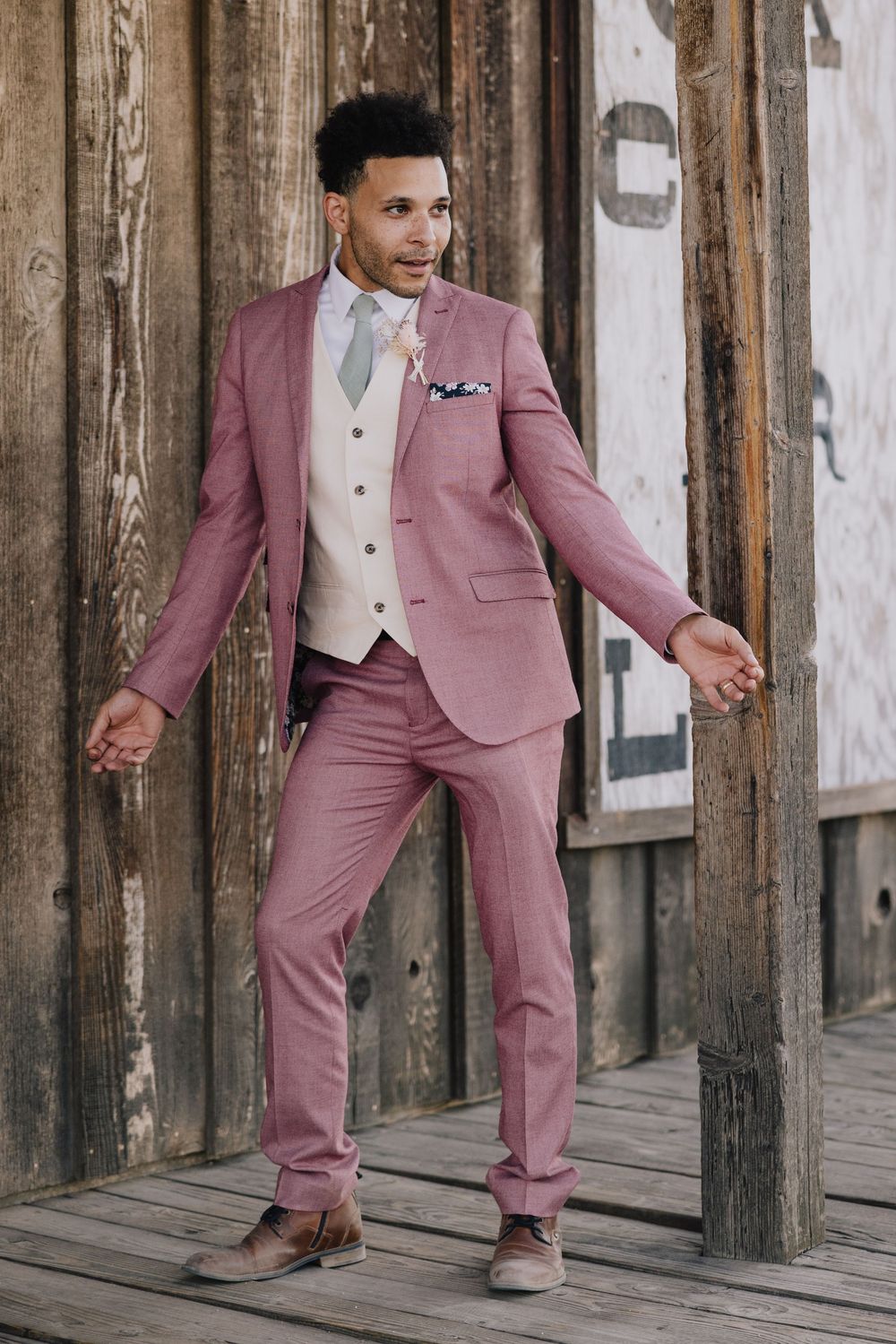 Man in pink suit and cream vest poses confidently outdoors near rustic wooden building.