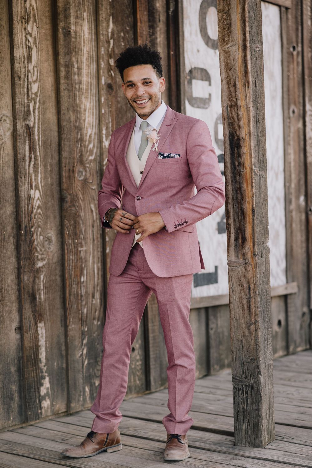 Smiling groom in pink suit with tie and pocket square poses against weathered wooden wall.