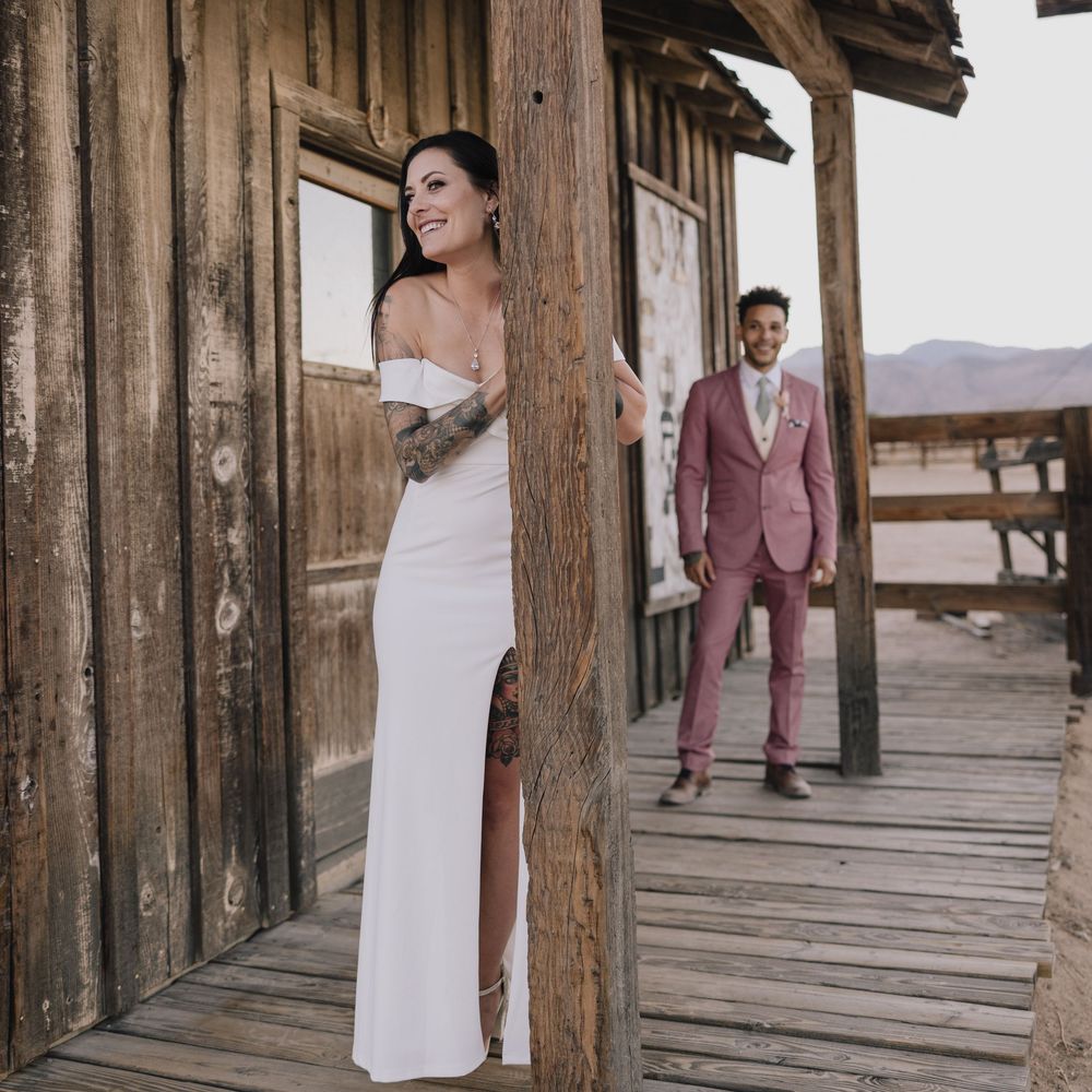 A bride in a white off-shoulder gown and groom in a pink suit pose at a rustic wooden building.