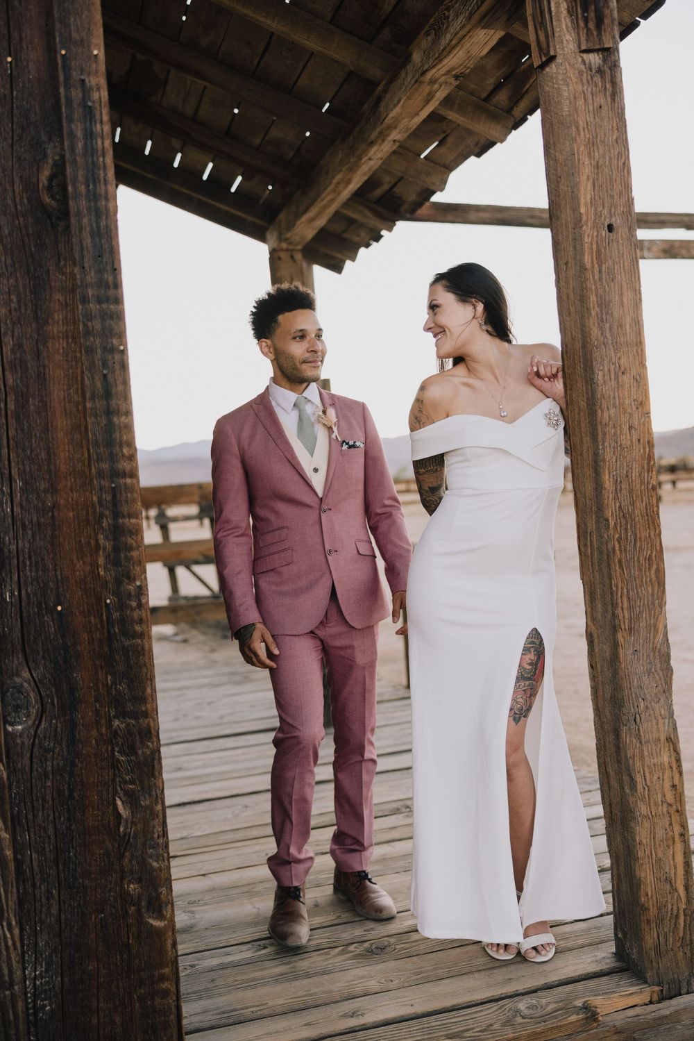 Couple in pink suit and white off-shoulder gown on rustic wooden deck.