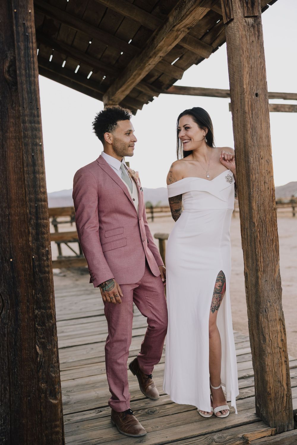 Bride and groom in pink suit standing under rustic wooden structure on weathered deck.