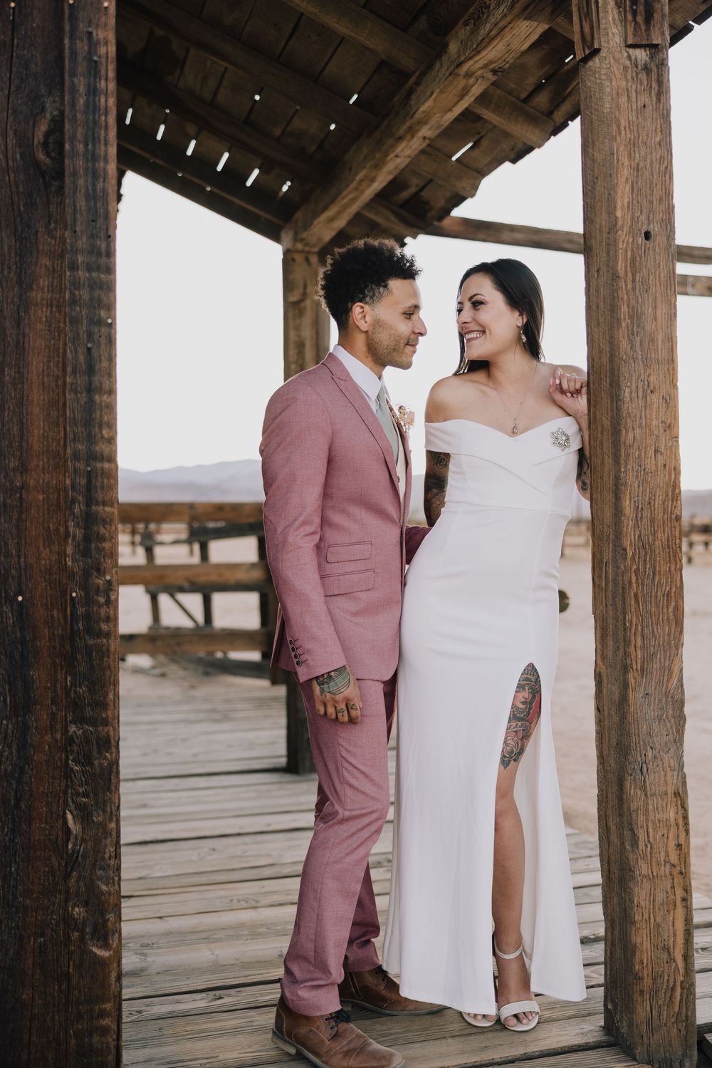 Newlyweds share a joyful moment under wooden structure, groom in pink suit, bride in white gown.