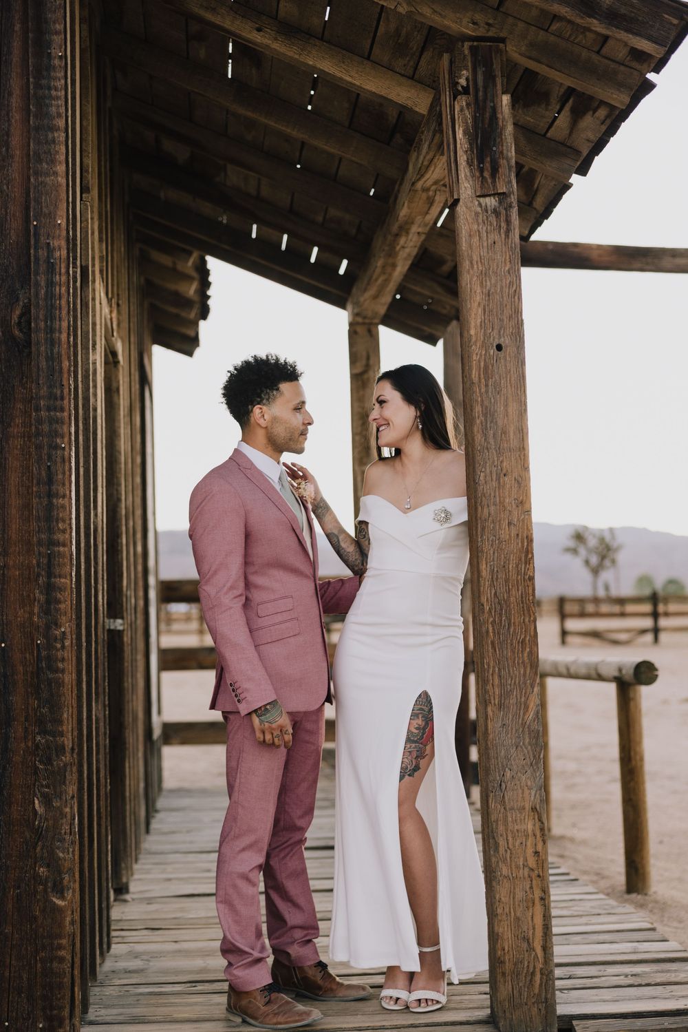 Bride and groom in white dress and pink suit pose on rustic wooden porch at desert wedding venue.
