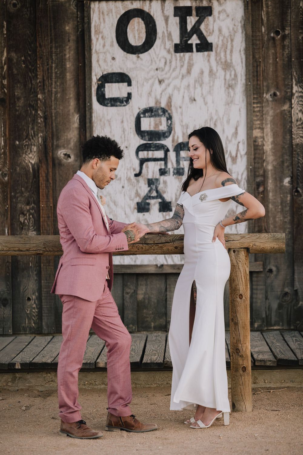 Couple in pink suit and white wedding dress pose before rustic wooden wall with vintage OK Corral sign.
