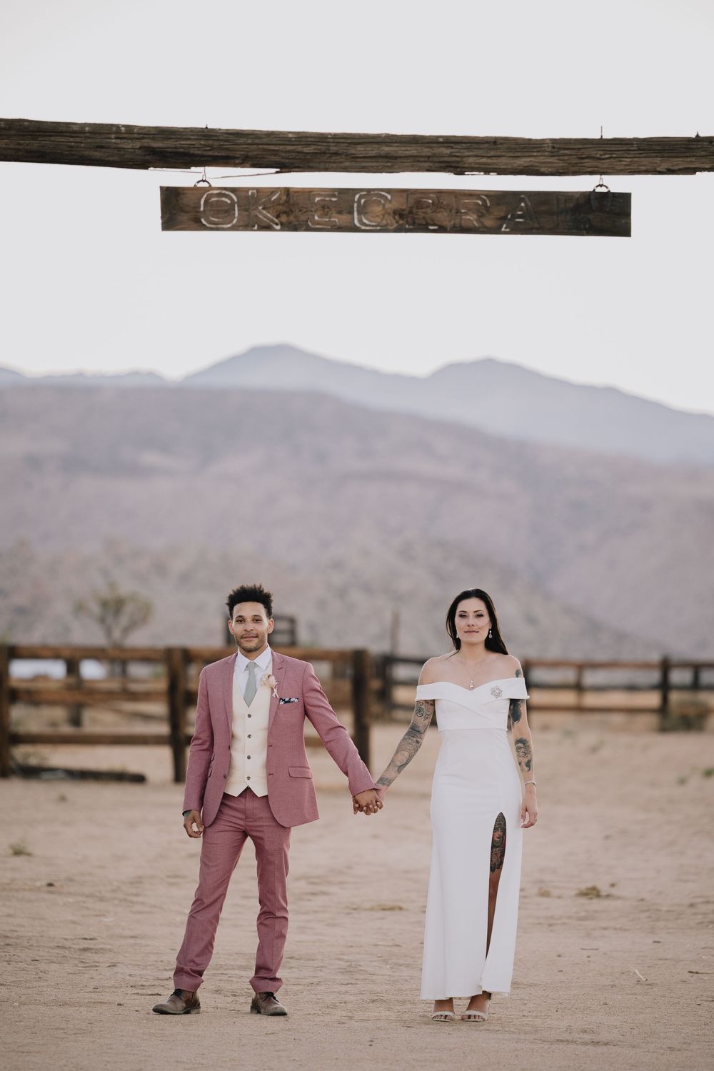 Couple in pink suit and white gown hold hands at desert wedding venue with mountain backdrop.