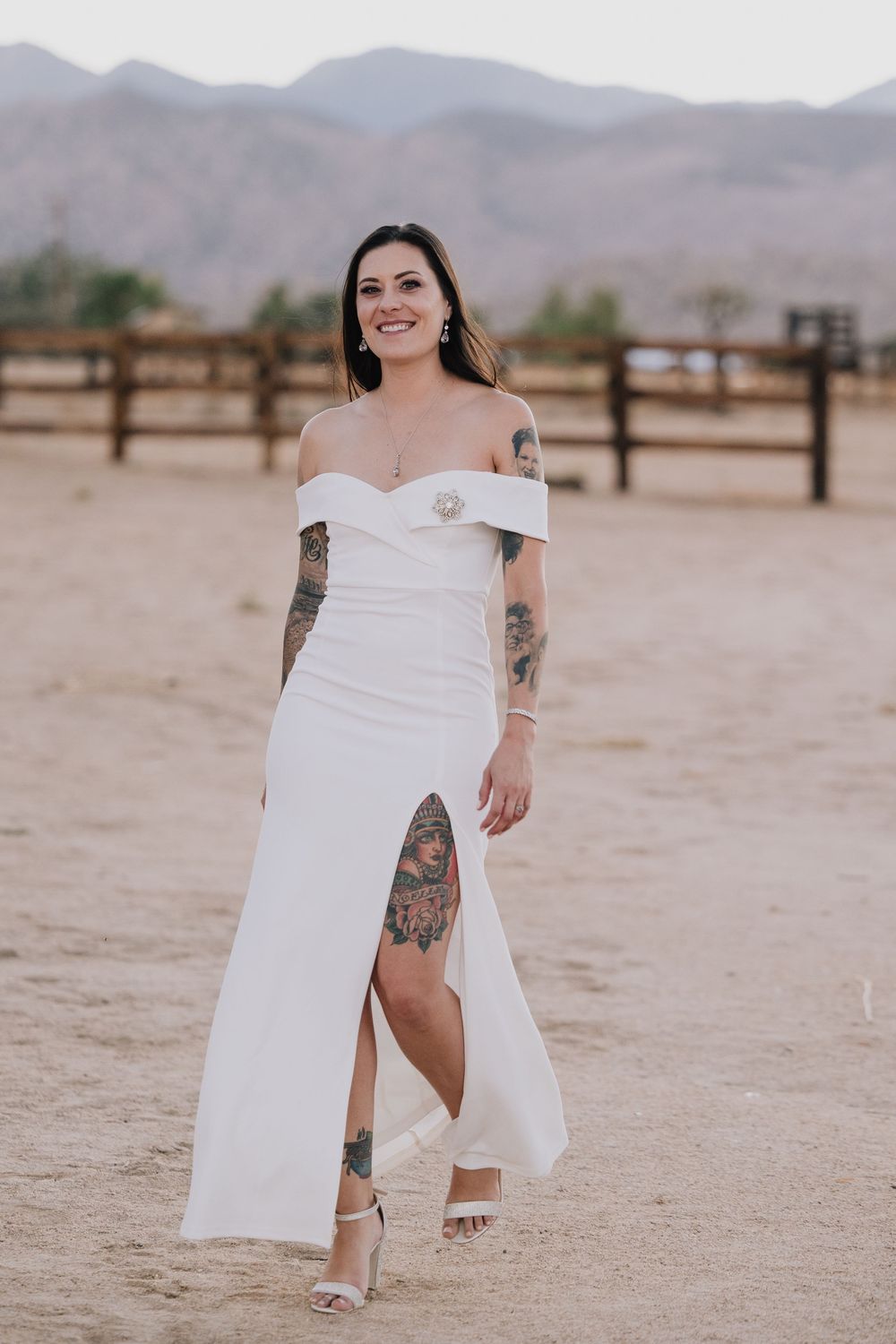 Tattooed bride in off-shoulder white gown with slit walks through desert venue with mountains behind.