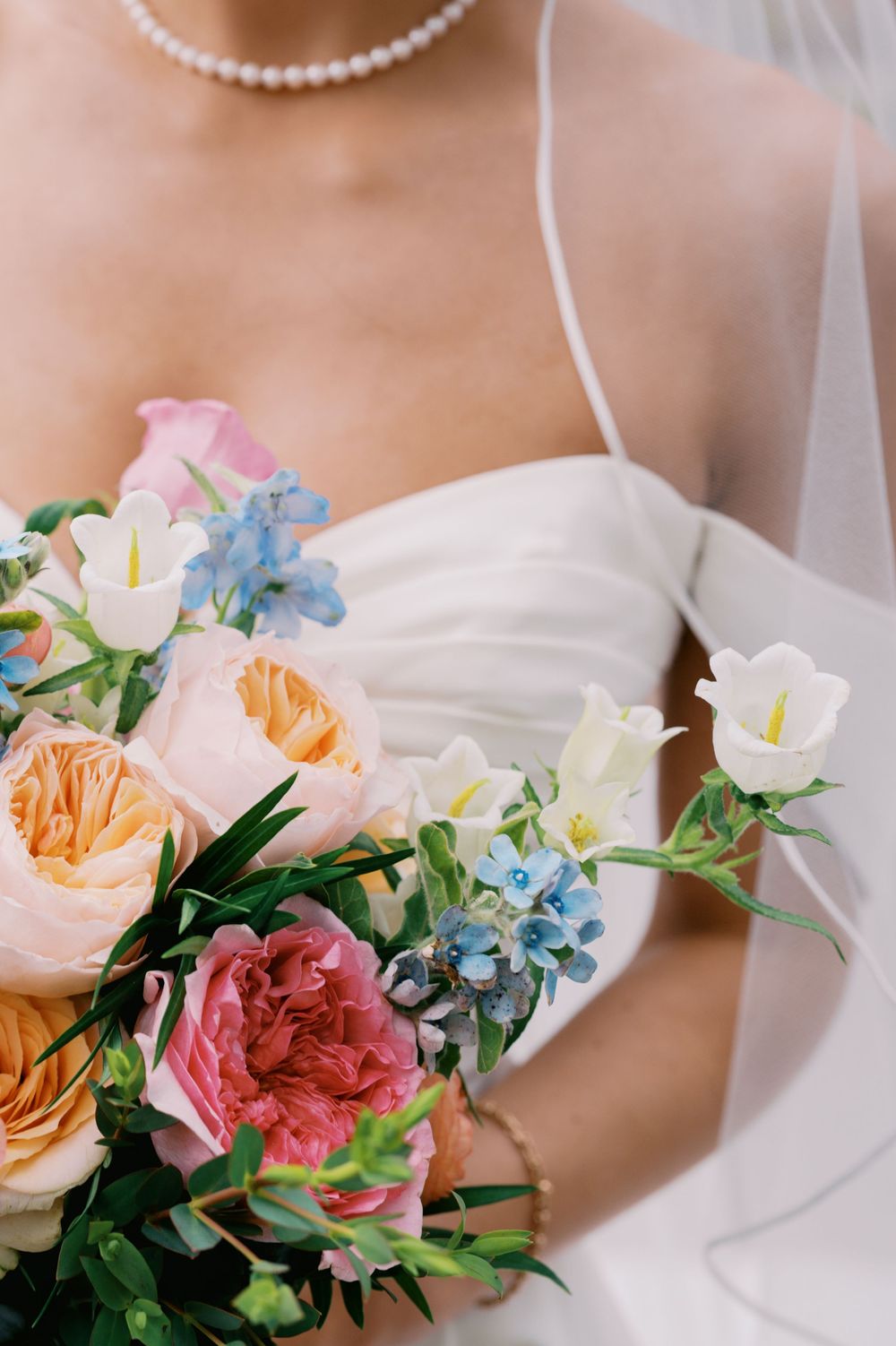 A pearl necklace and delicate floral bouquet with pink roses and blue flowers rest against white fabric.