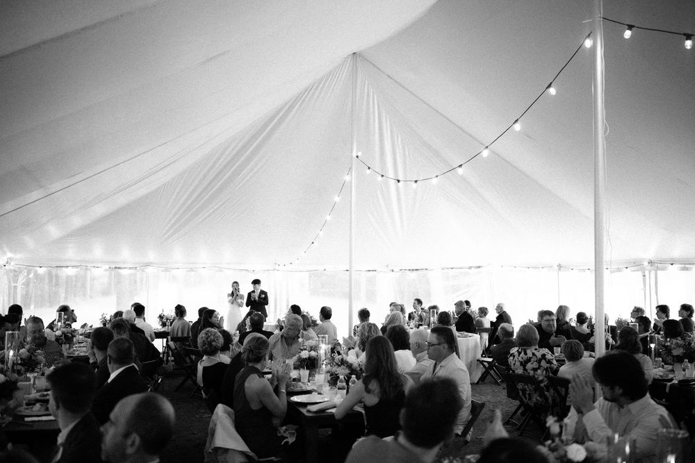 A black and white panoramic view of a crowded reception under a large tent with string lights overhead.