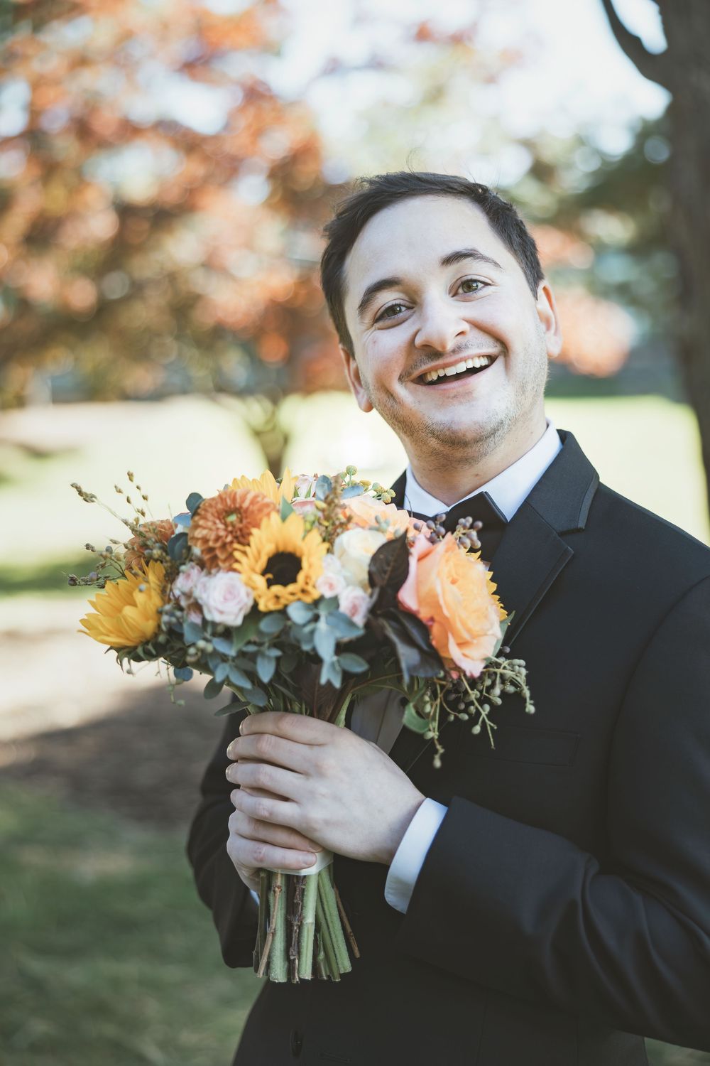 A joyful person in a black suit holds a vibrant autumn bouquet with sunflowers and peach blooms.