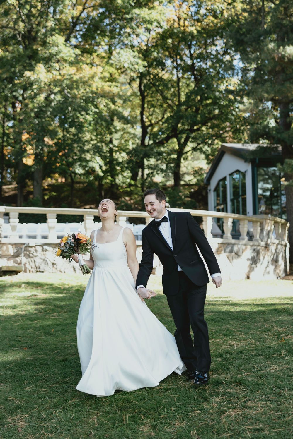 A joyful couple in wedding attire dance and twirl together on a sunny lawn with white fencing in the background.