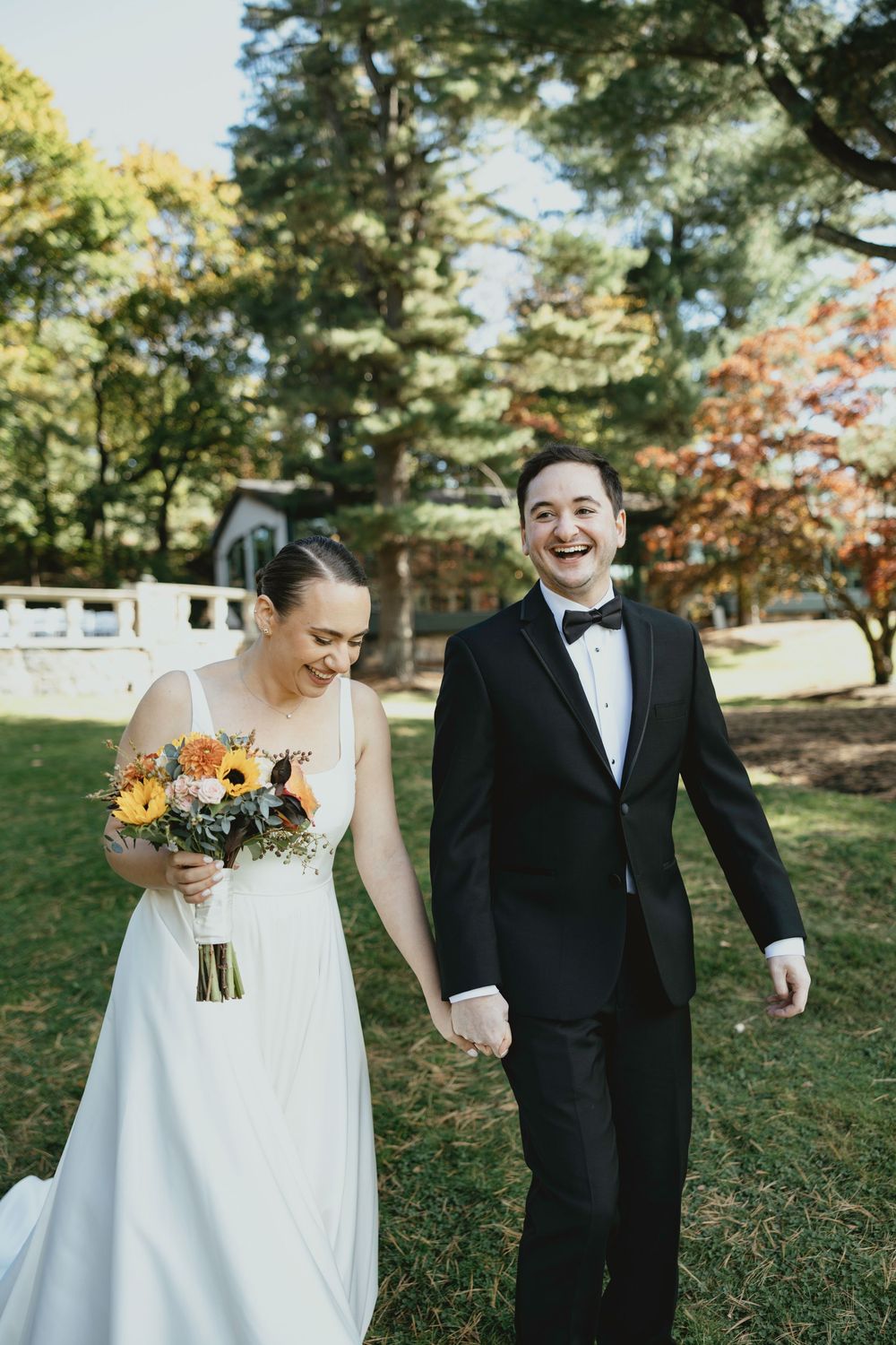 A couple shares a joyful moment walking together in a sunny outdoor setting with autumn trees in the background.