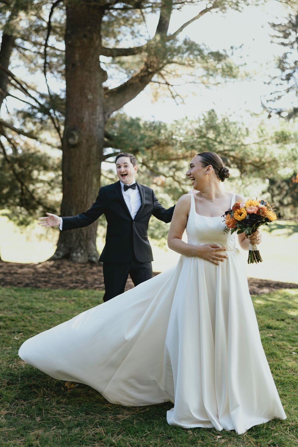 A joyful wedding moment outdoors with a bride in flowing white dress holding orange flowers and groom in black tuxedo.