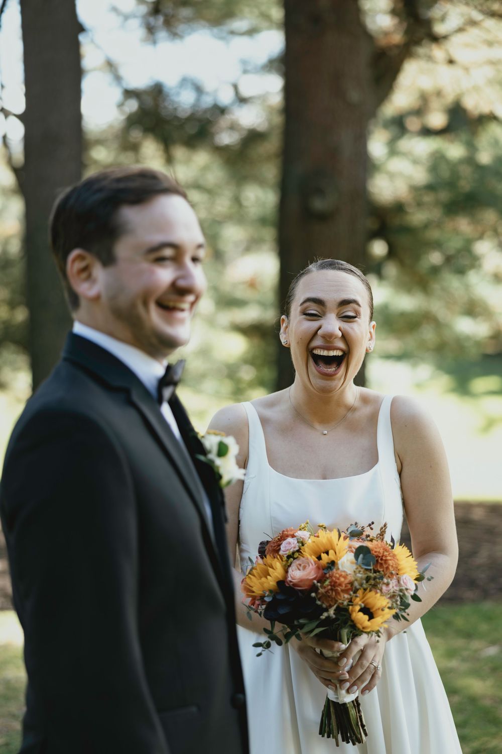 A couple laughs together during their wedding photoshoot in a wooded outdoor setting with sunlit trees.