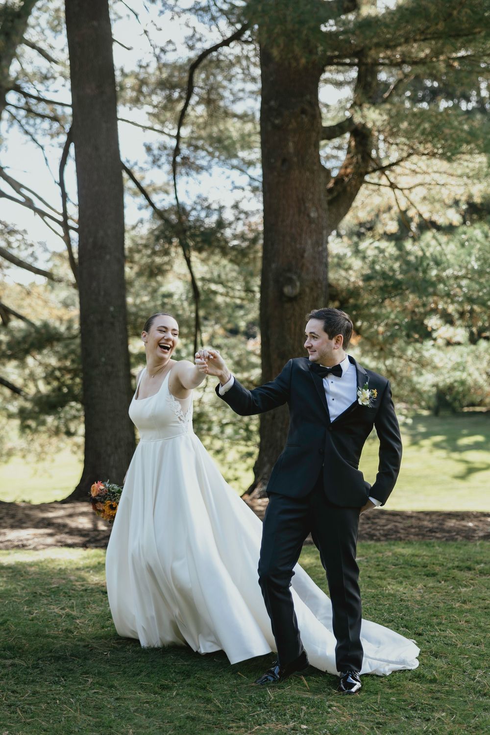 Newlywed couple dances playfully in a sunlit forest setting surrounded by tall pine trees.