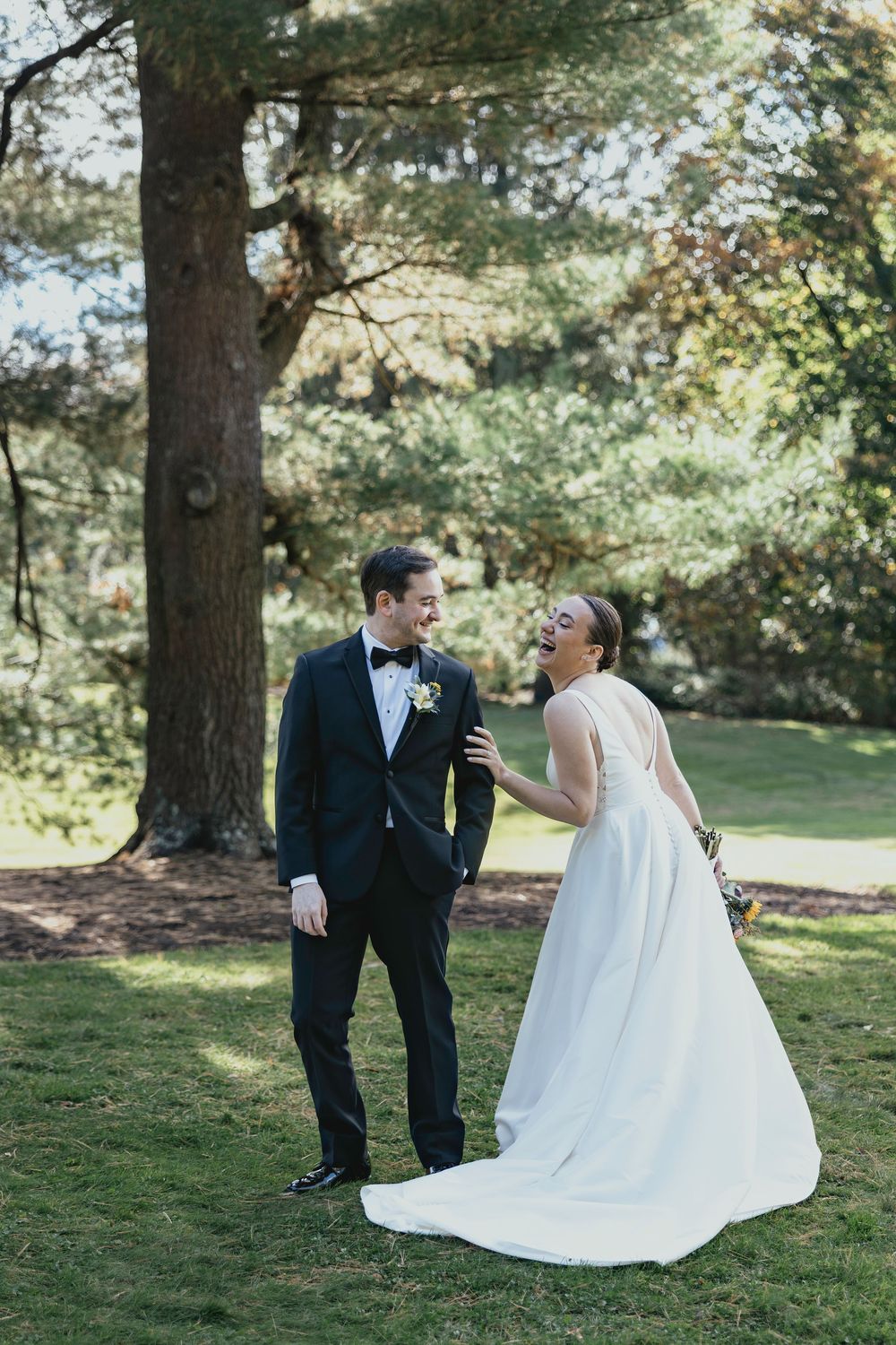 A couple shares playful moments in formal wedding attire while posing on grass under sunlit trees.