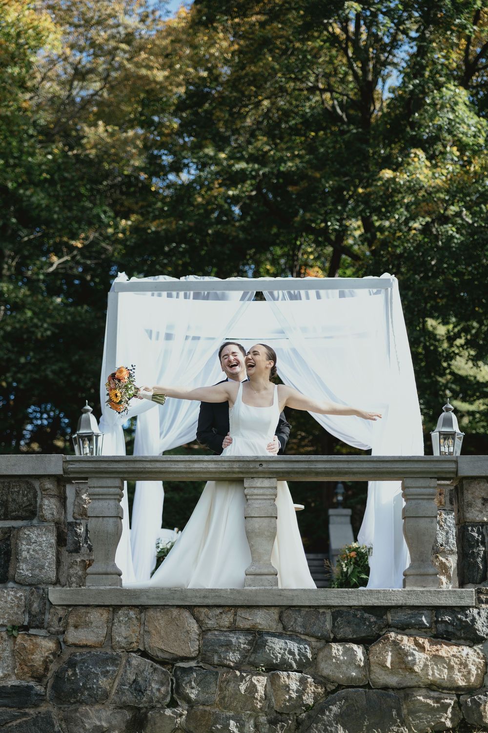 Wedding couple poses behind white drapes on stone wall in romantic outdoor garden setting.