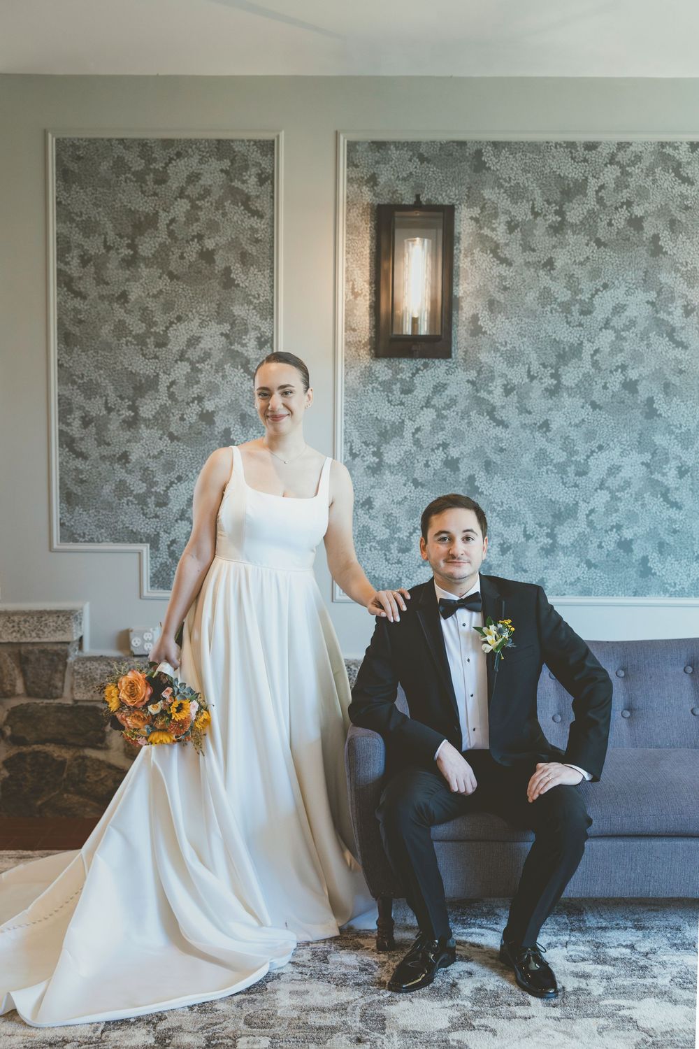 A wedding couple lounges playfully on a gray sofa against floral wallpaper in a bright room.