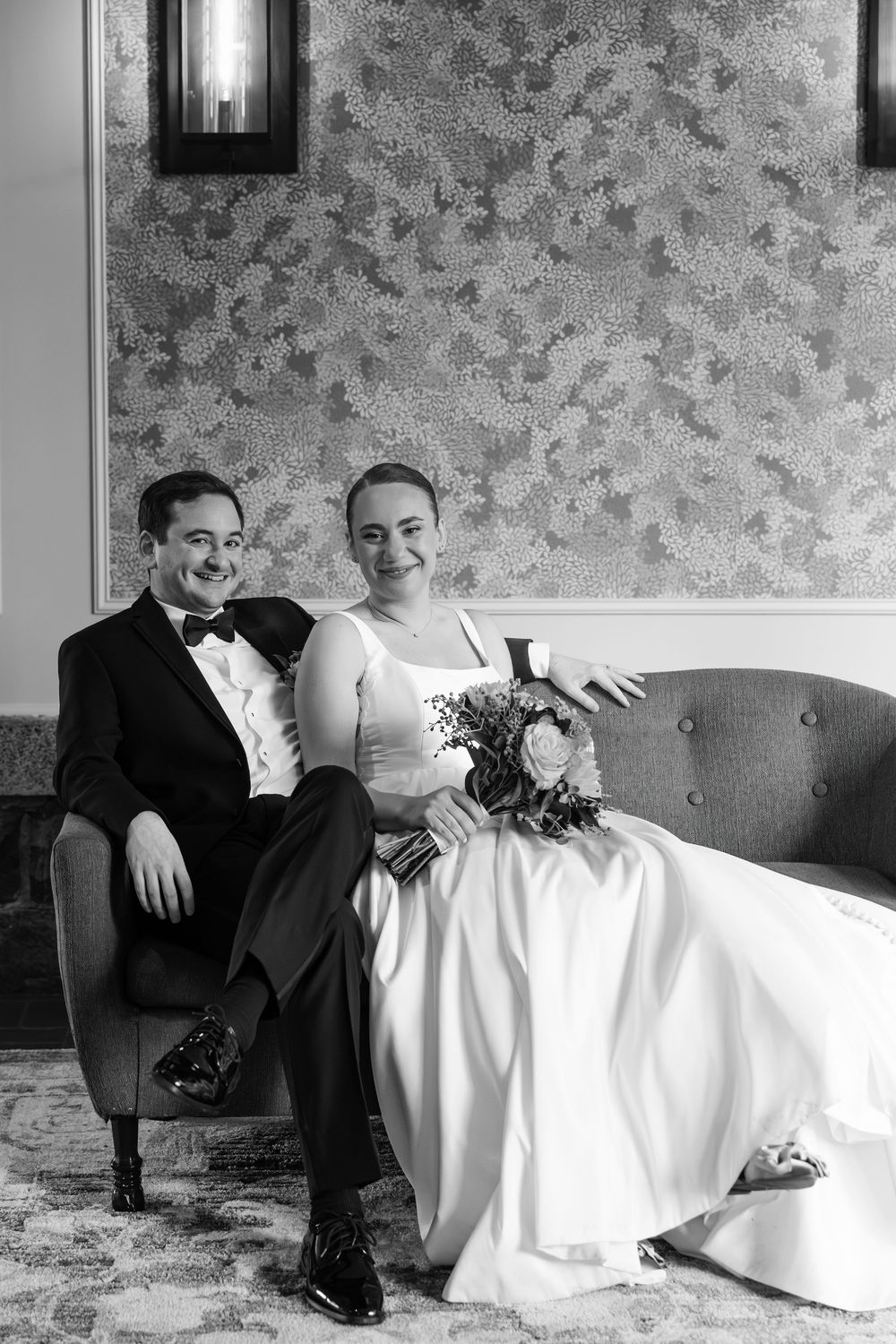 Black and white wedding photo of couple relaxing on vintage sofa with floral wallpaper backdrop.