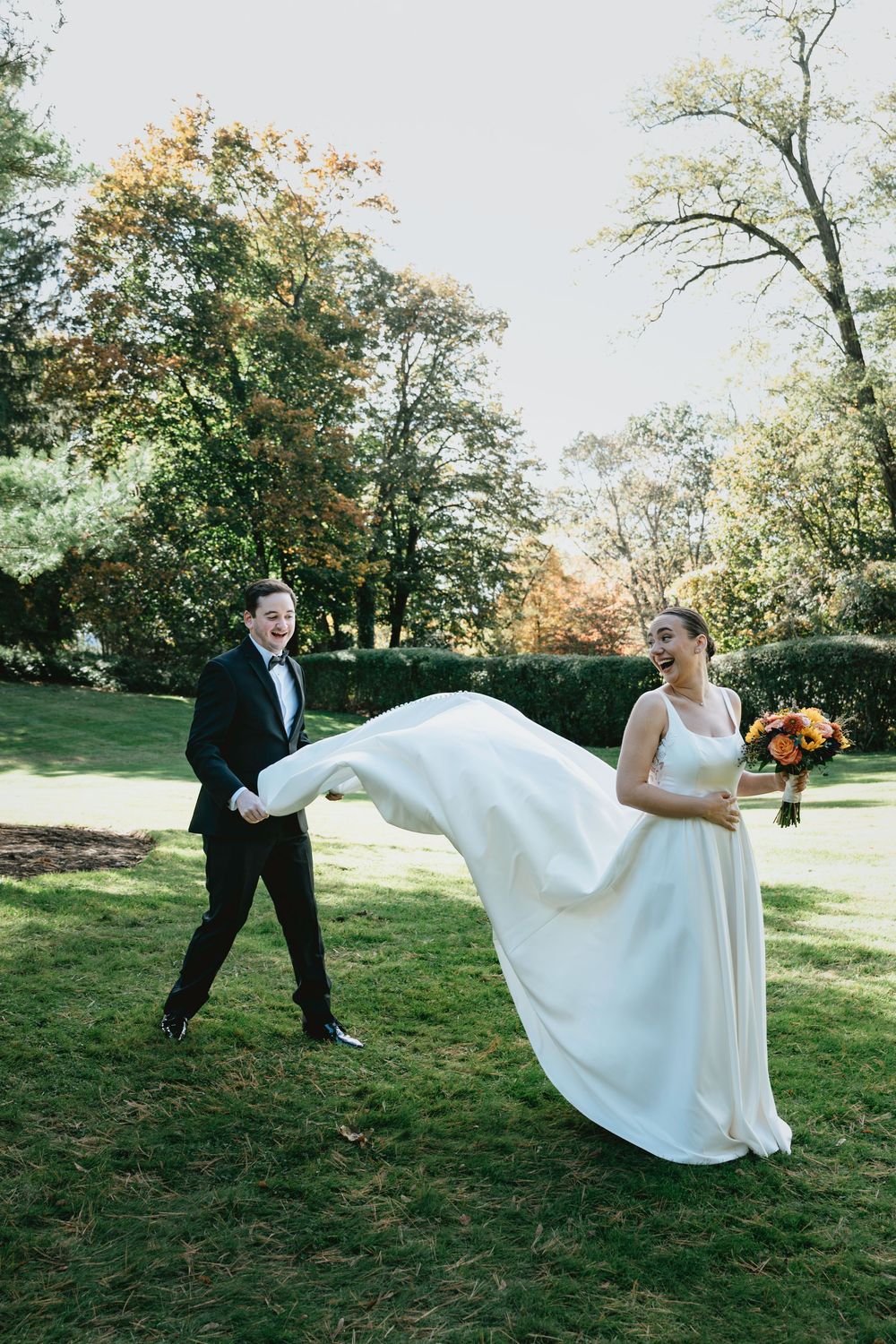 A fun wedding portrait of a couple playing with the bride's dress flowing in the wind on a sunny lawn.