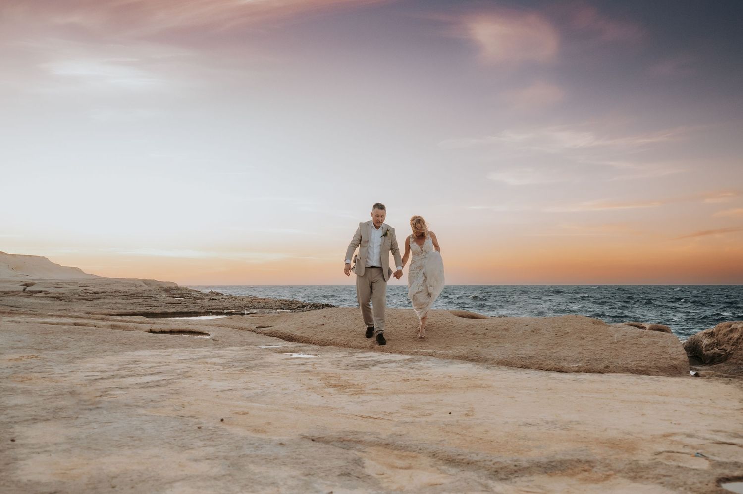 A couple walks hand in hand along a sandy beach at sunset with warm golden light illuminating the shoreline.