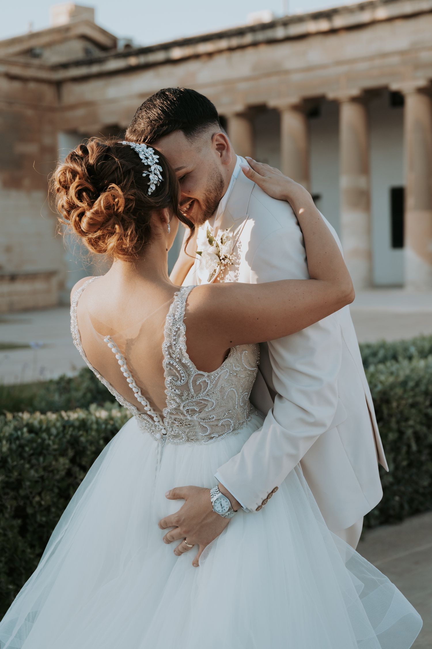 Romantic wedding couple dancing together in elegant white dress and suit against stone building backdrop.