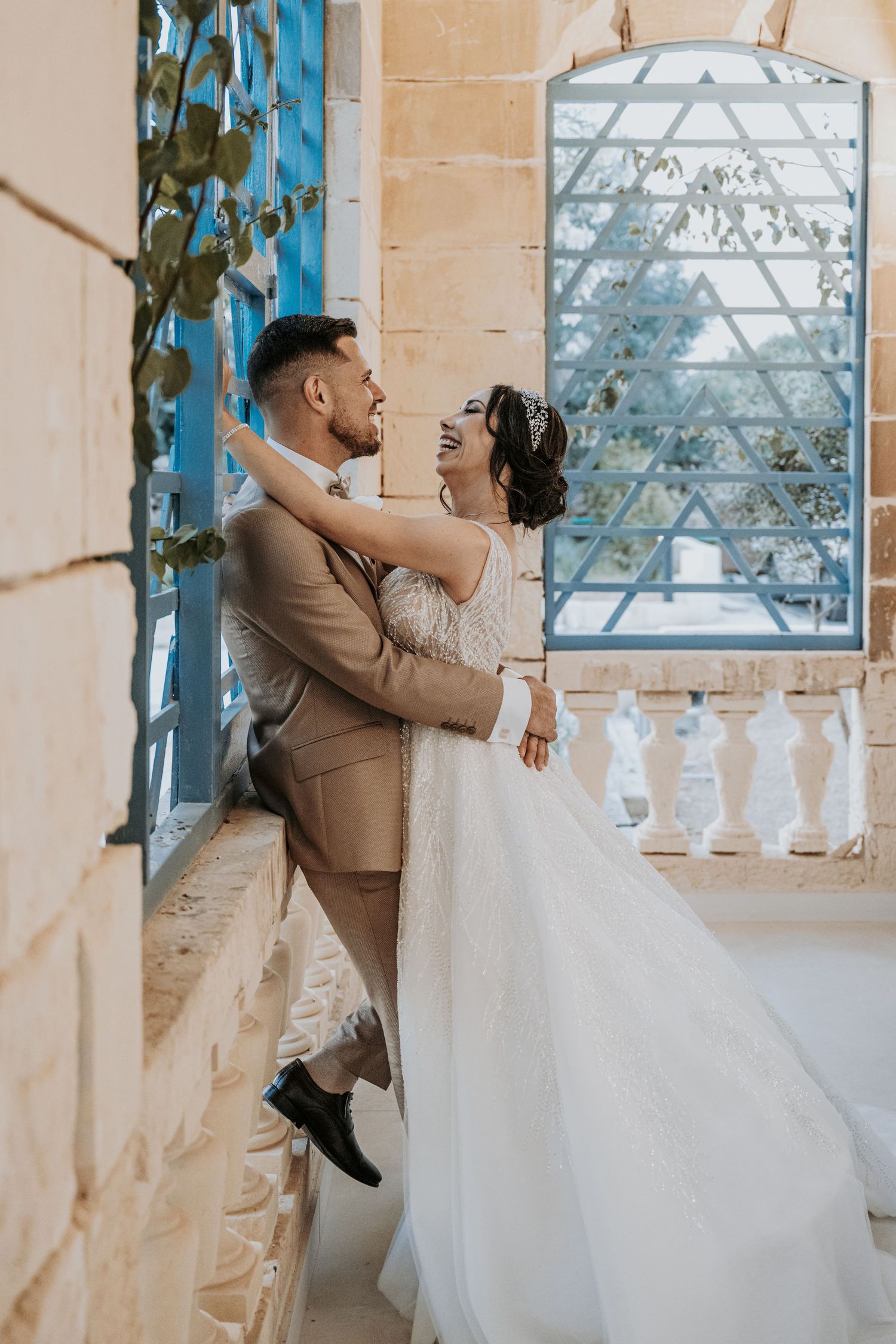 A couple in wedding attire dances playfully near a window in a light-filled room with beige walls and blue trim.