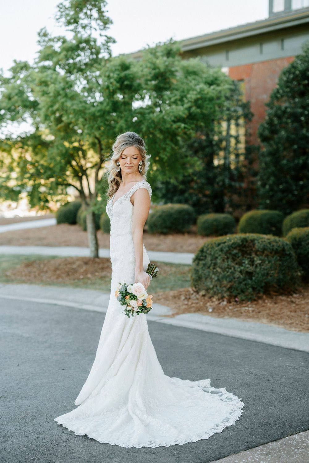 A bride in a flowing lace wedding gown with a train stands on a driveway surrounded by manicured landscaping at sunset.