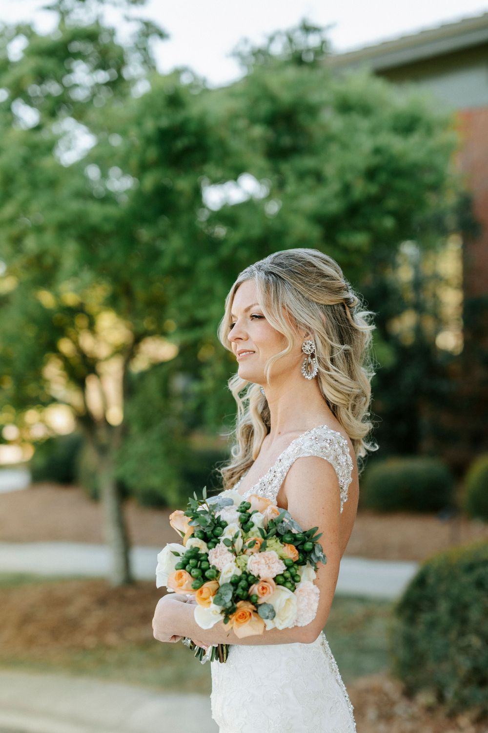 A bride in a lace wedding dress smiles while holding a beautiful bouquet of peach and white flowers outdoors.