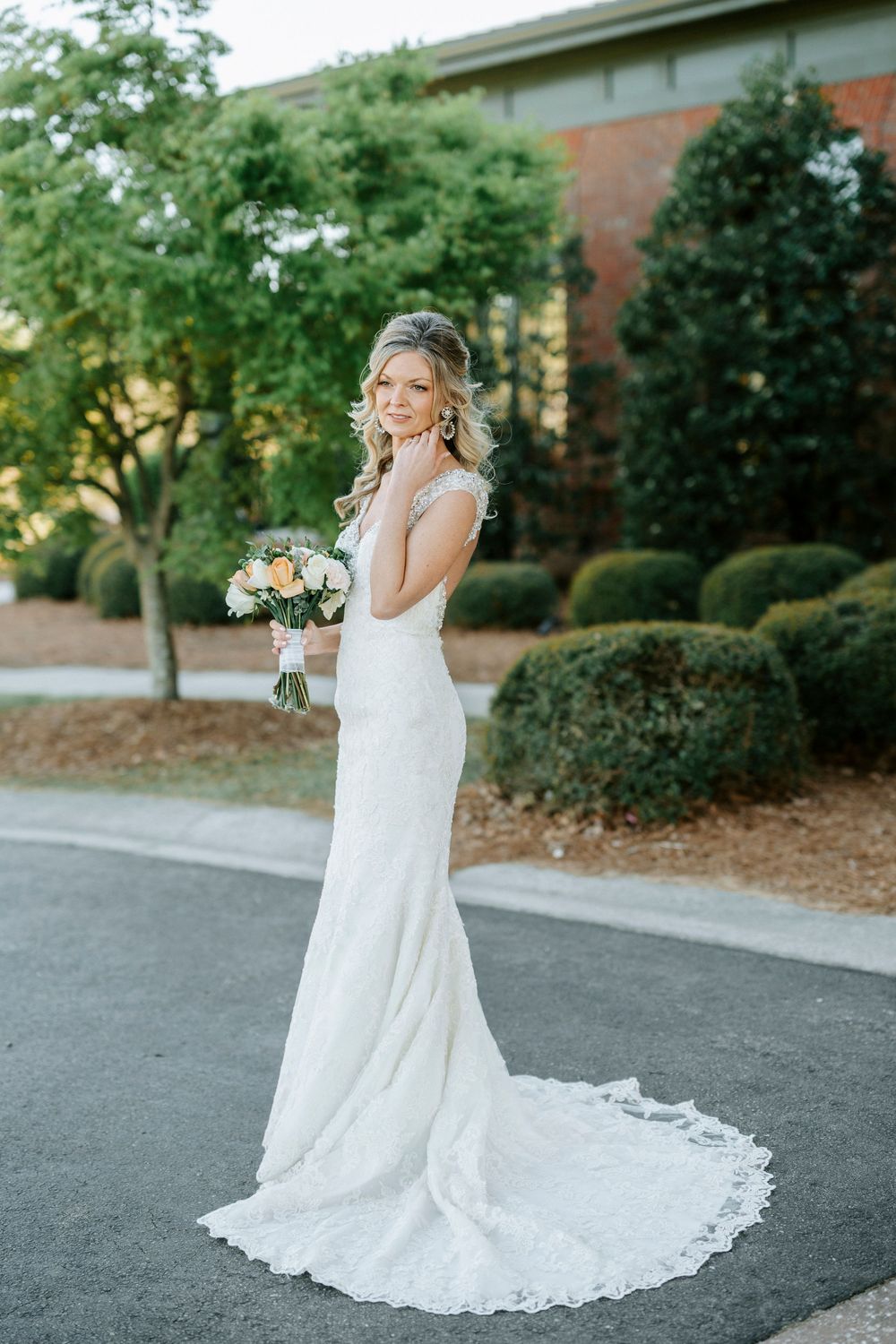 A bride in a flowing white lace wedding gown with train stands outdoors holding a bouquet on a paved walkway.