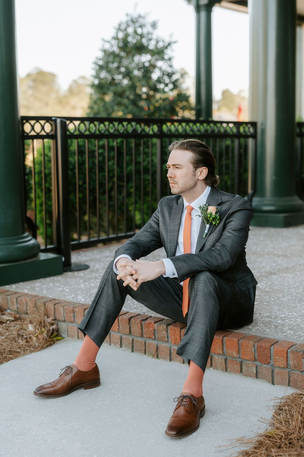 Professional wearing a gray suit and orange tie sits casually on brick steps with brown leather shoes.